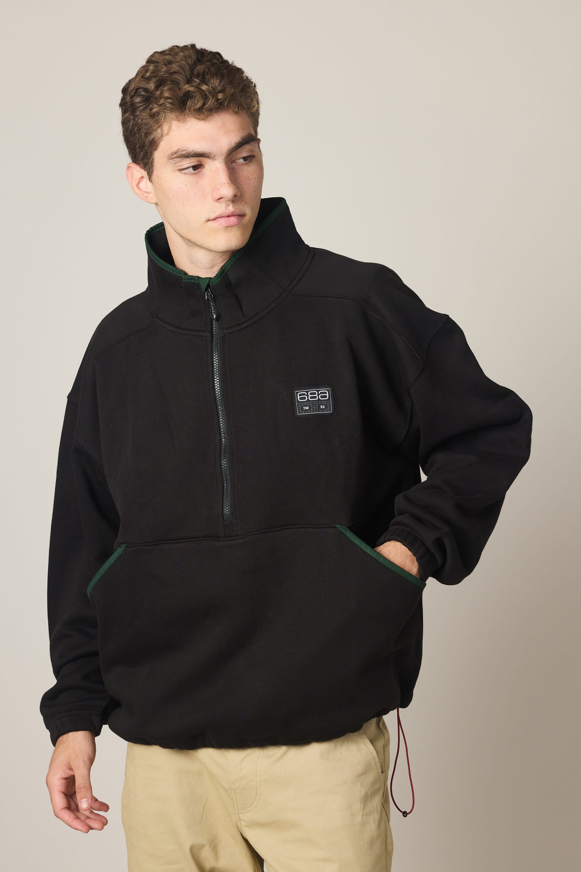 A young man stands against a plain background, looking to his left with one hand in his pocket. He's wearing the 686 Men's Burnout Anorak—a black waterproof jacket with a small chest patch—paired with light-colored pants.