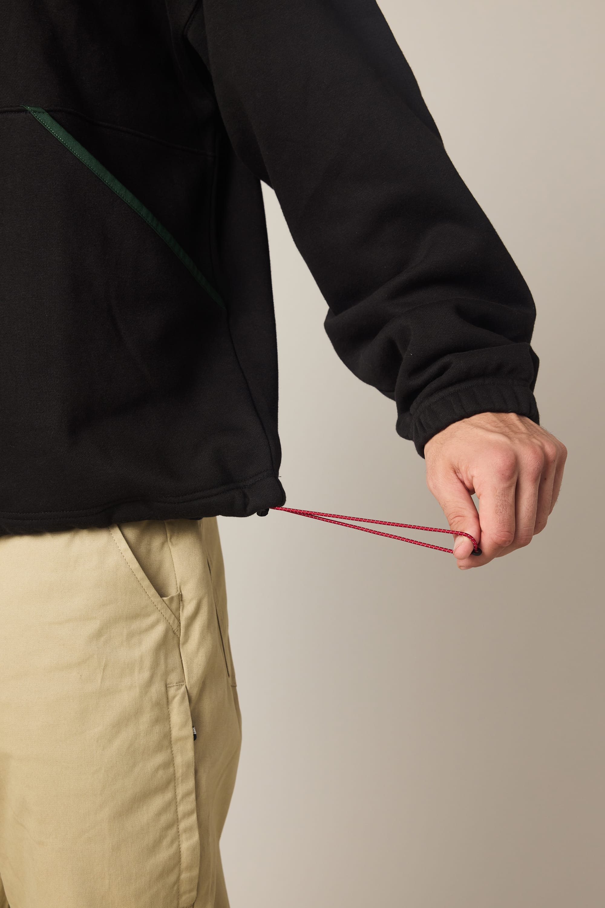 A person in the 686 Men's Burnout Anorak, a black waterproof jacket, and beige pants is pulling the red drawstring at the hem to tighten it. Only their hand, arm, part of their torso, and legs are shown.