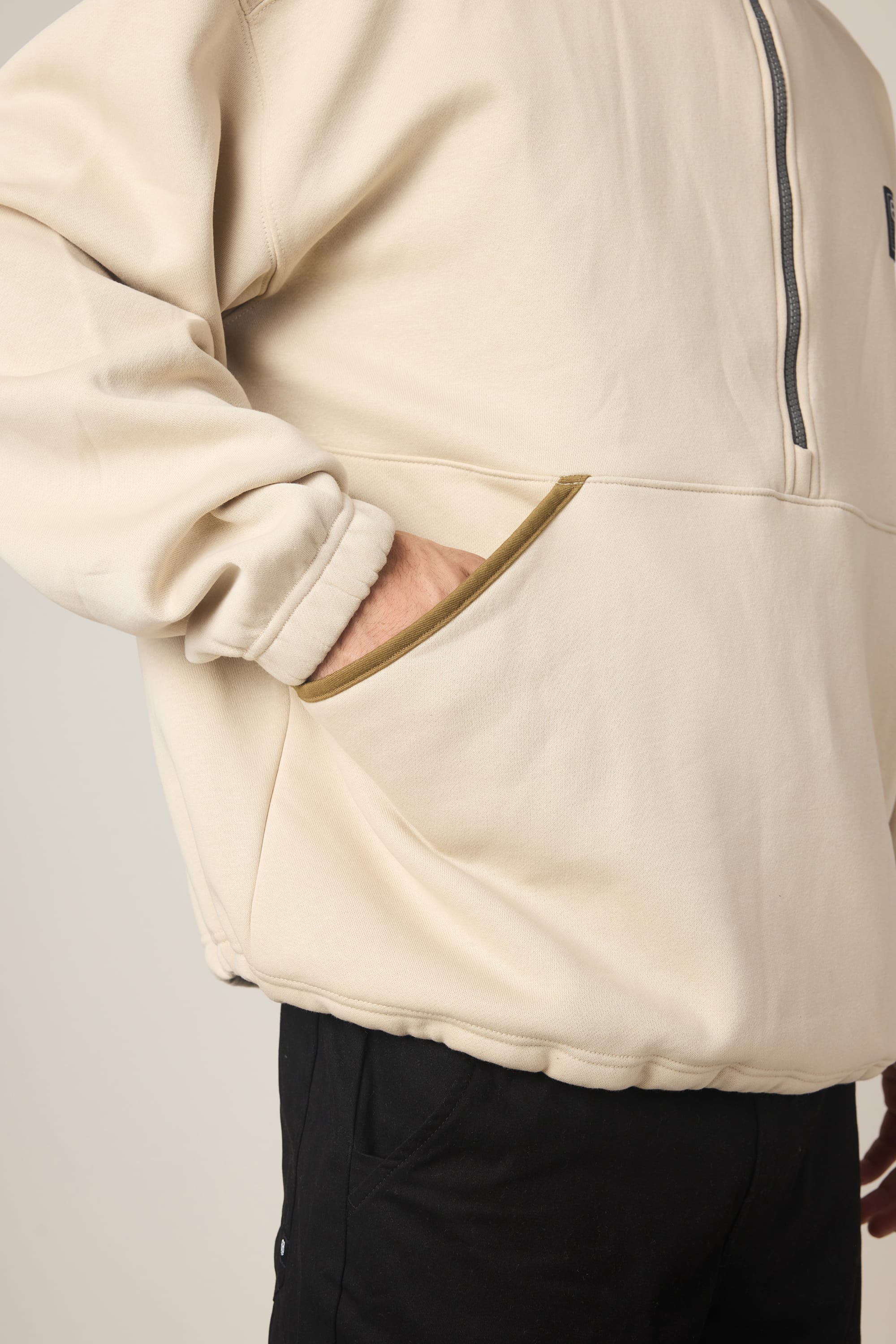 A person wearing the 686 Men's Burnout Anorak in beige, featuring a zip-up front and one hand in the pocket, paired with black pants. Shown from the waist up against a neutral background.