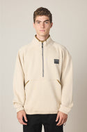 A young man with short curly hair wears the 686 Men's Burnout Anorak, a light beige quarter-zip pullover jacket with a front pocket and black chest patch, standing facing forward against a plain light background.