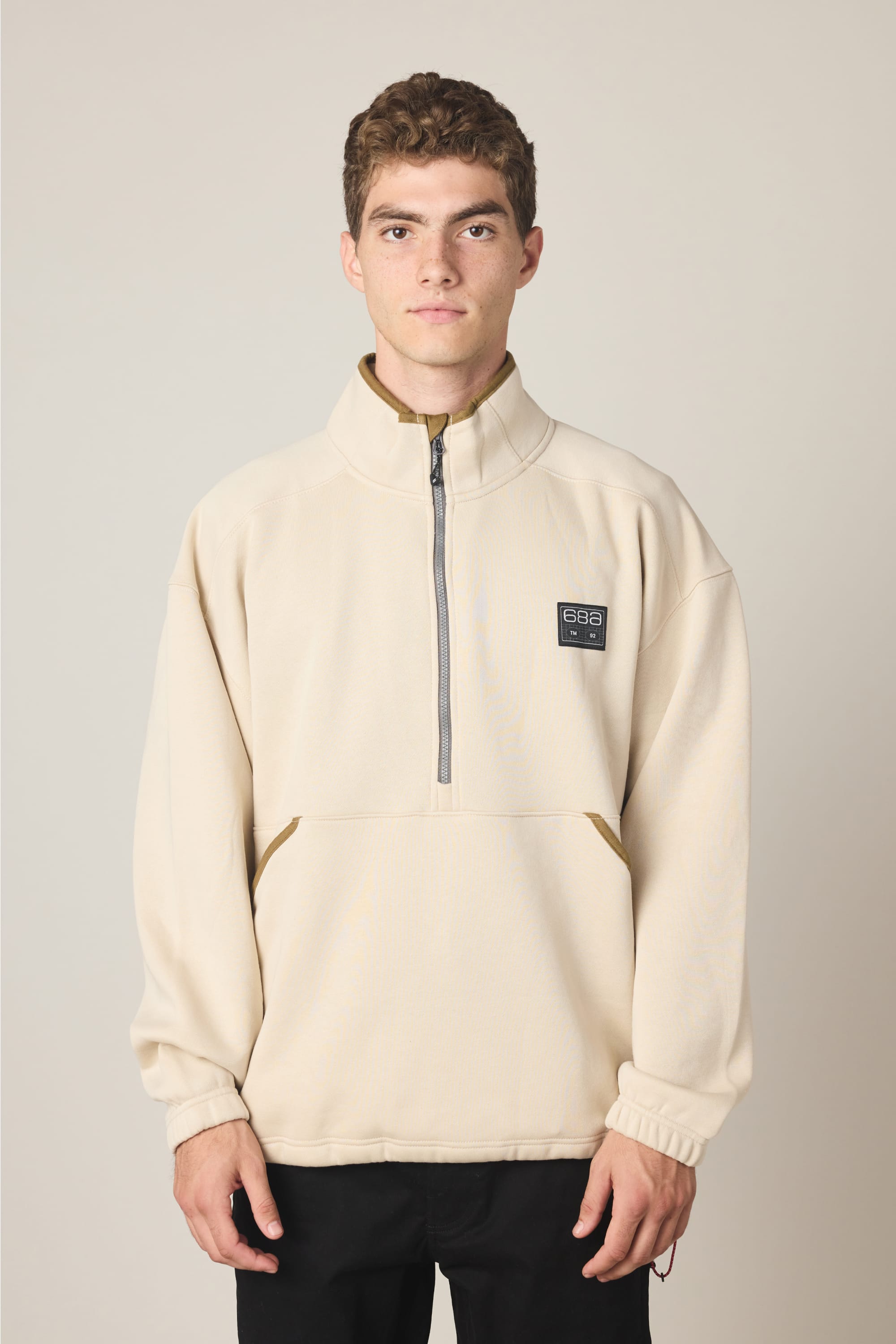 A young man with short curly hair wears the 686 Men's Burnout Anorak, a light beige quarter-zip pullover jacket with a front pocket and black chest patch, standing facing forward against a plain light background.