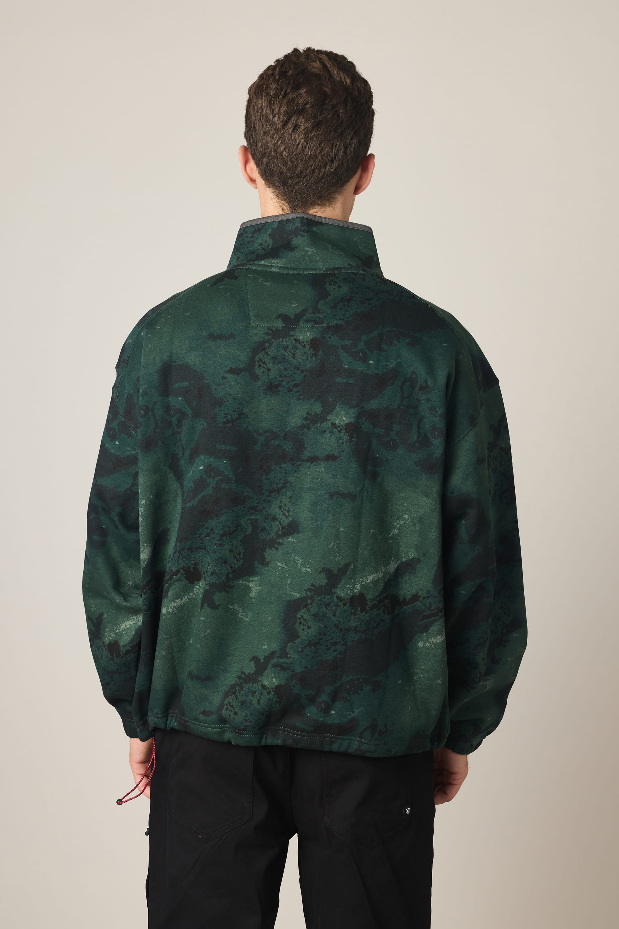A person with short brown hair is shown from behind, wearing the 686 Men's Burnout Anorak in a green and black camouflage pattern with black pants against a light background.