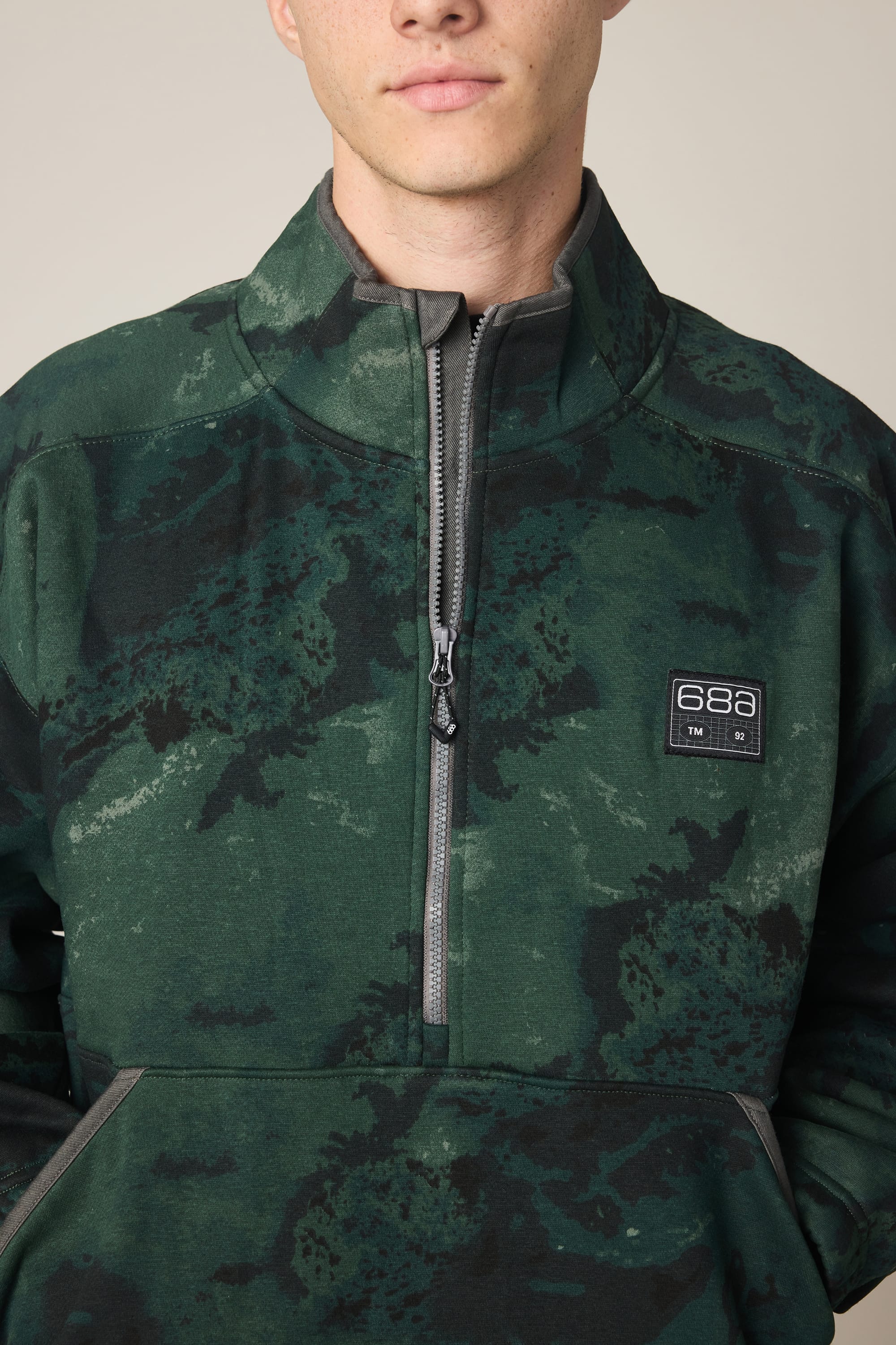 A person wearing the 686 Men's Burnout Anorak—a green, camouflage-patterned woven jacket with a half-zip and a 686 logo patch on the chest—stands against a neutral background, with only the lower part of their face visible.