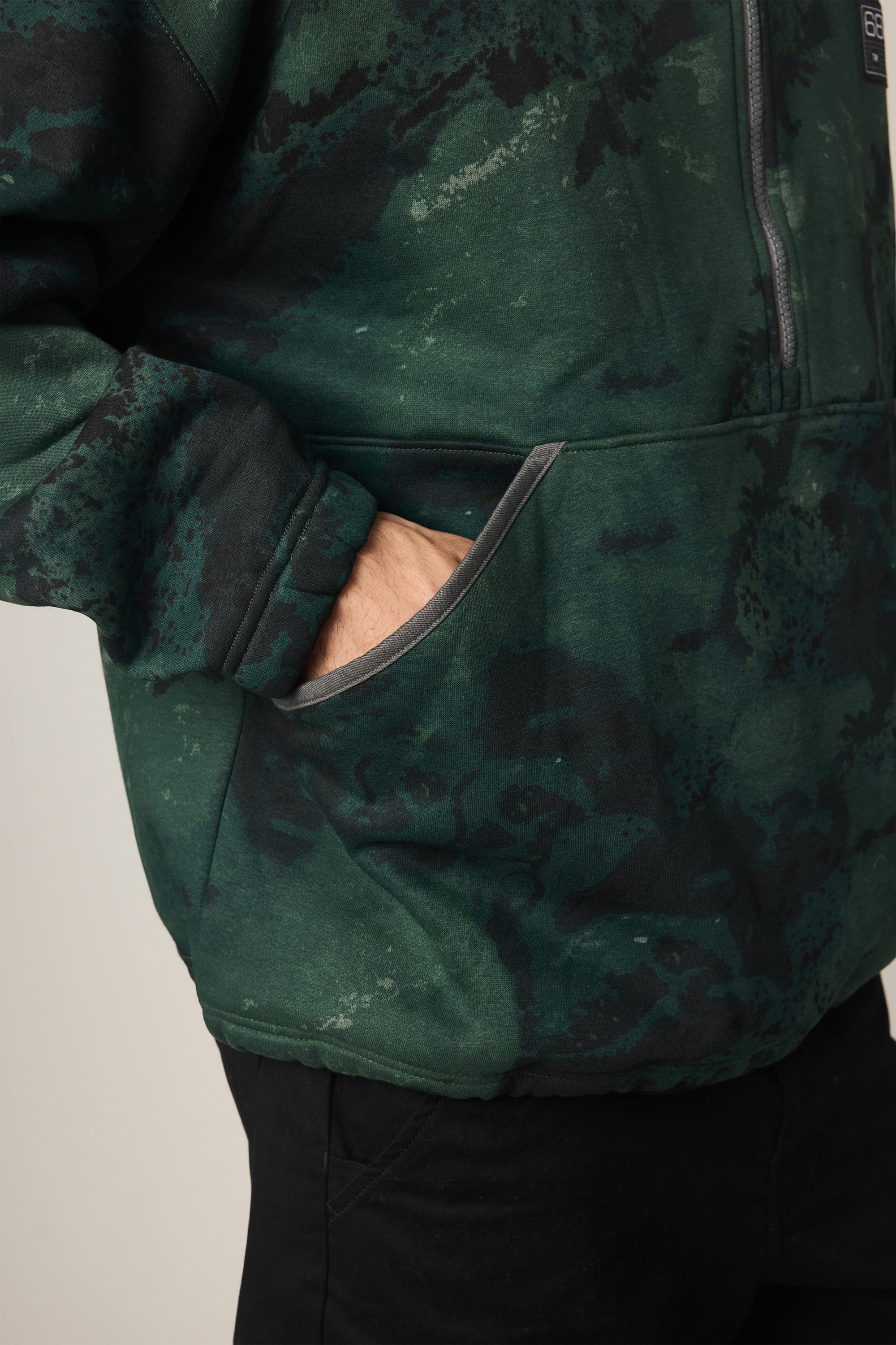 Wearing the 686 Men's Burnout Anorak by 686, a person is shown from the torso up with one hand in the front pocket, highlighting the jacket's dark green and black patterned design.