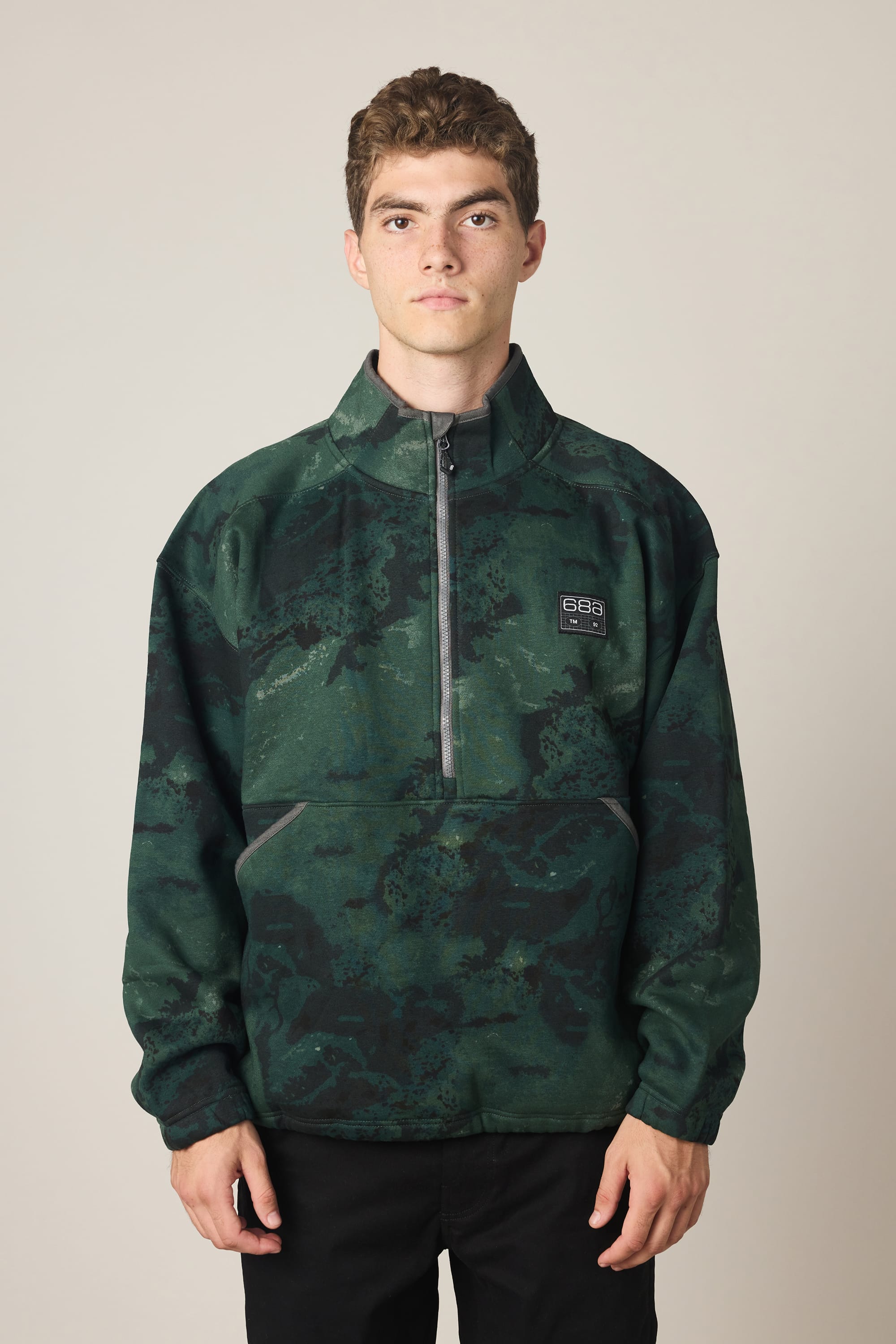 A young man with short brown hair wears the 686 Men's Burnout Anorak, a green and black camouflage woven jacket with a half-zip and a 686 logo on the left chest, standing against a plain light background.