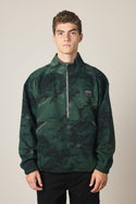 A young man with short curly hair wears the 686 Men's Burnout Anorak, a dark green and black camouflage jacket with a small chest logo by 686, standing against a plain light-colored background.