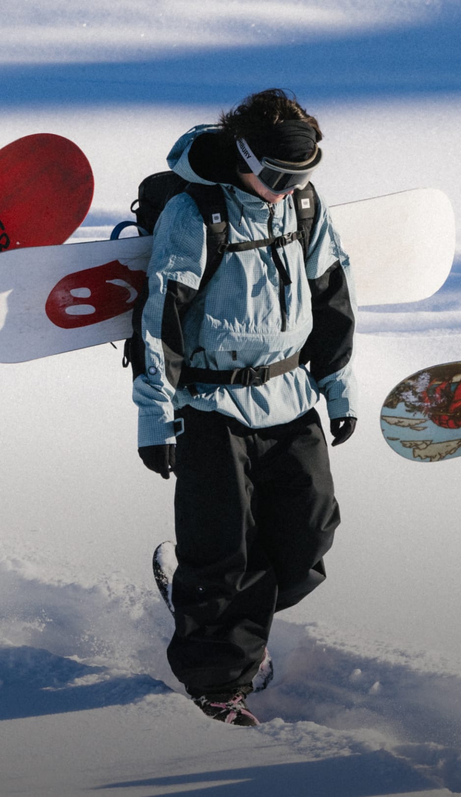 A person in snowboarding gear walks through snow carrying a snowboard on their backpack, with snowboards and snowy terrain visible in the background.