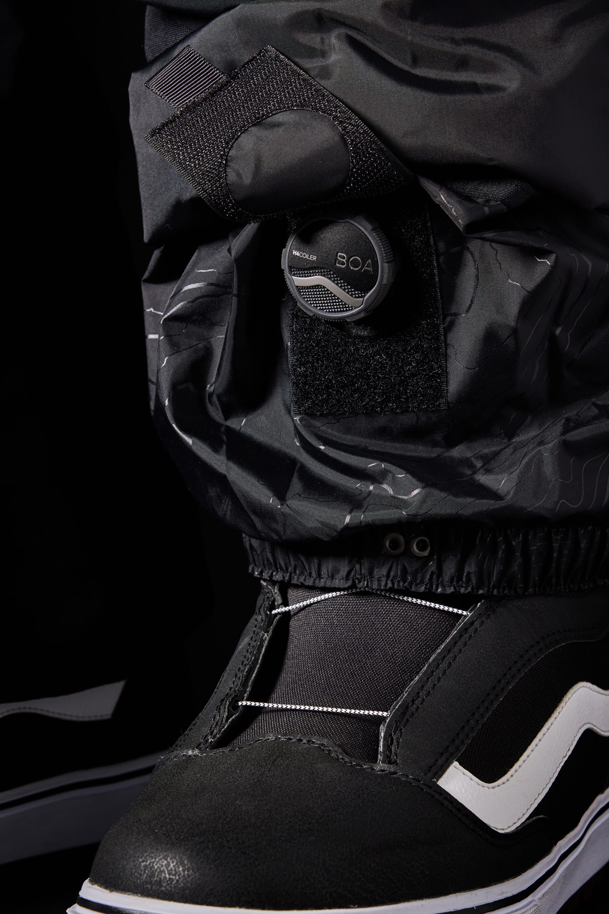 Close-up of the 686 Men's SMARTY® 3-in-1 Cargo Pant over a black snowboard boot with a BOA dial, reflective details, white stripe, and three wear options via a removable sweatpant liner.