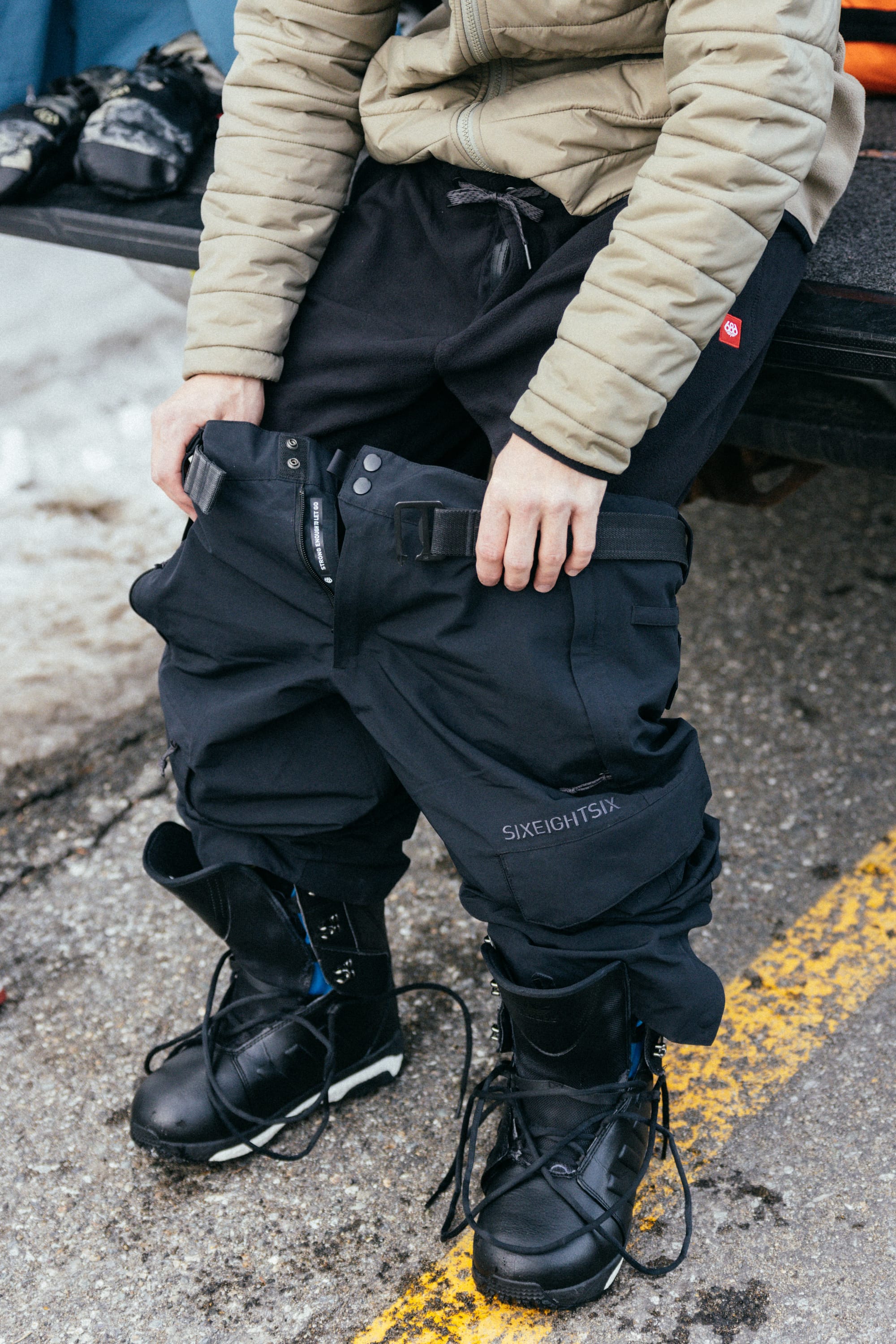 A person sitting on a vehicle tailgate puts on 686 Men's SMARTY® 3-in-1 Cargo Pants with a removable liner from 686 and black snow boots, wearing a beige jacket. Snow is visible on the ground nearby.