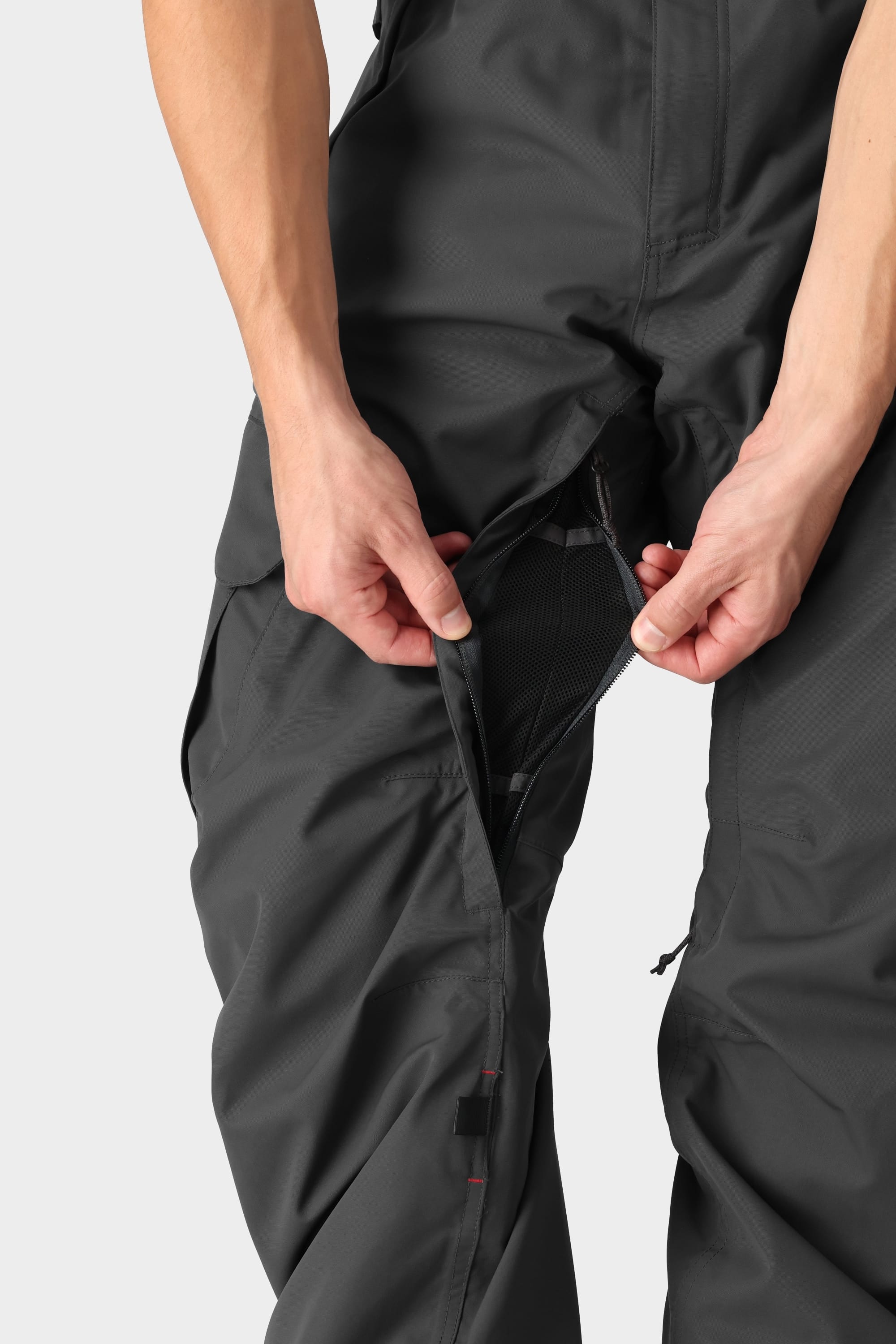 A person unzips a thigh vent on the 686 Men's SMARTY® 3-in-1 Cargo Pant, revealing mesh lining. The hands and pants are in focus, showcasing three ways to wear the versatile 686 cargo pant against a plain background.