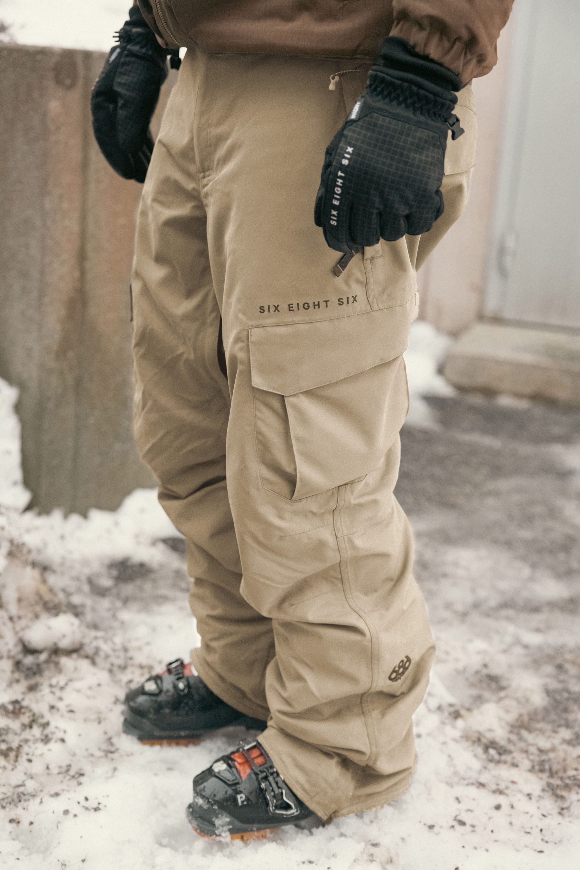 A person stands on snow wearing 686 Men’s SMARTY® 3-in-1 Cargo Pant in beige, paired with black gloves and black boots featuring orange accents. The pants have SIX EIGHT SIX branding and multiple pockets.