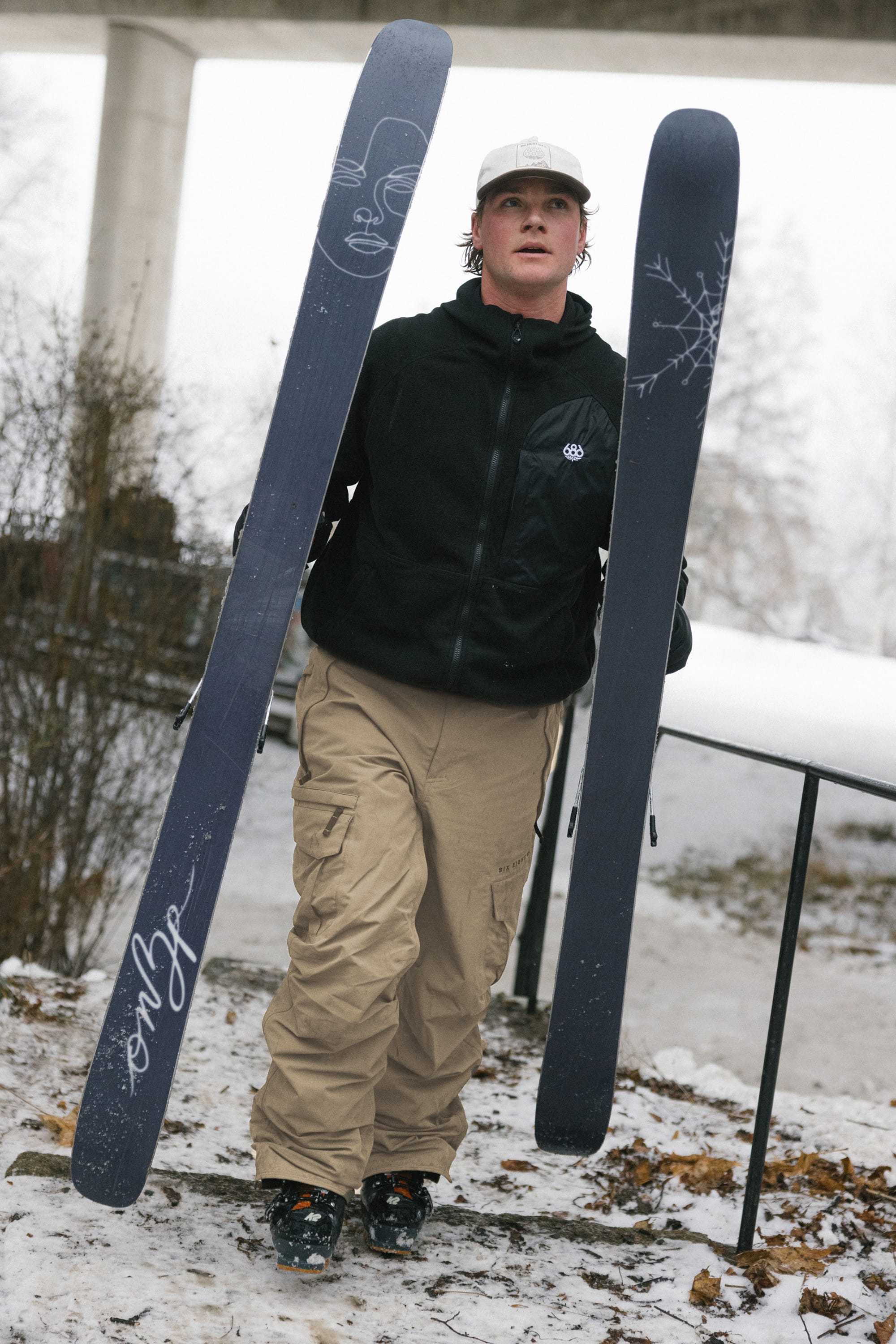 A person in a black jacket and 686 Men’s SMARTY® 3-in-1 Cargo Pant stands outside on a snowy day holding a pair of skis, with trees, snow, and a structure visible in the background.