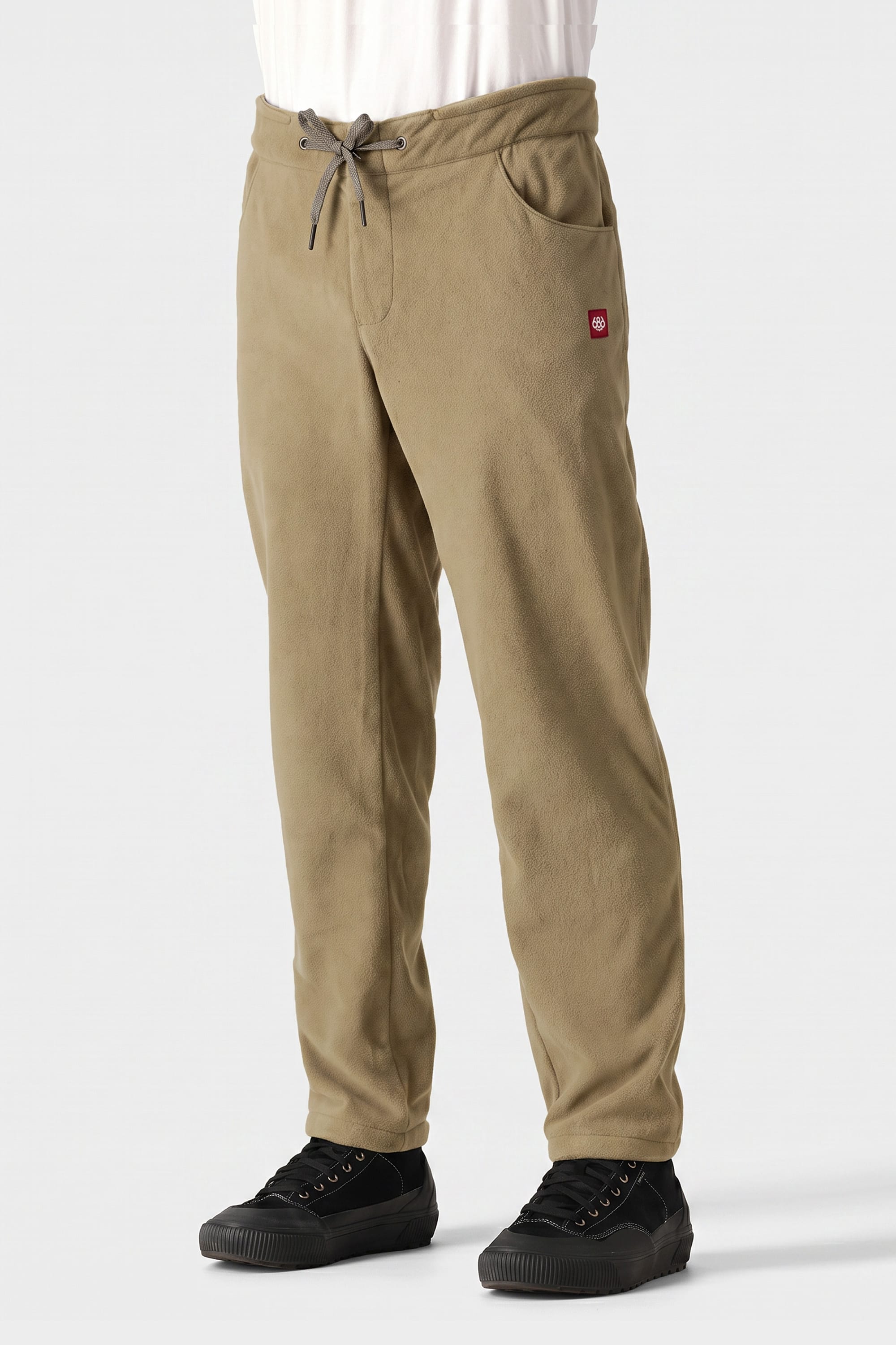 A person is wearing the 686 Men’s SMARTY® 3-in-1 Cargo Pant by 686, featuring side cargo pockets, a drawstring waist, and a small red logo patch on the left thigh. They are also wearing black sneakers, and the background is plain and light-colored.