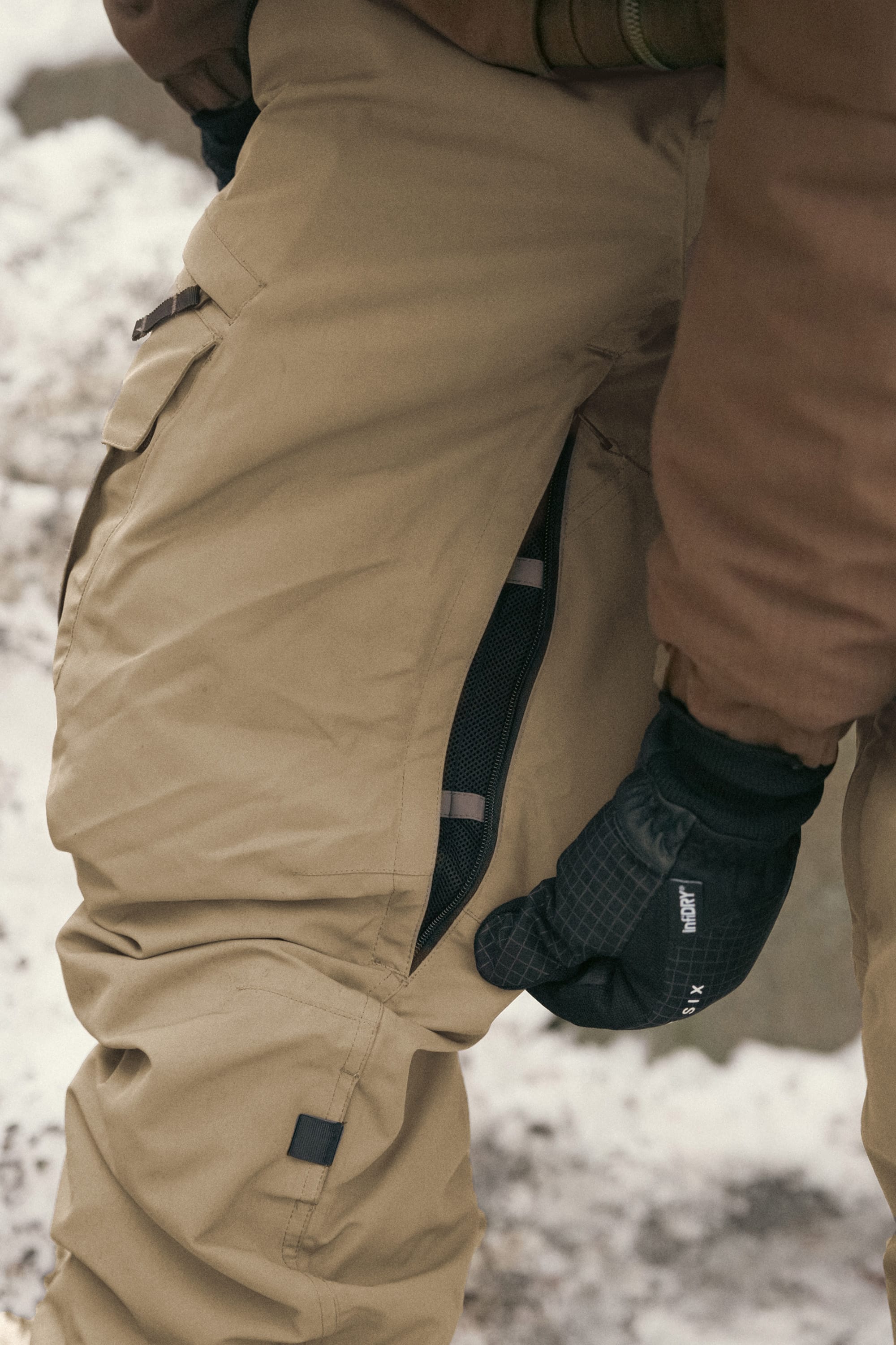 Wearing the 686 Men’s SMARTY® 3-in-1 Cargo Pant in tan and a brown jacket, a person opens the side zipper vent on the pants with a black-gloved hand as snow covers the background.