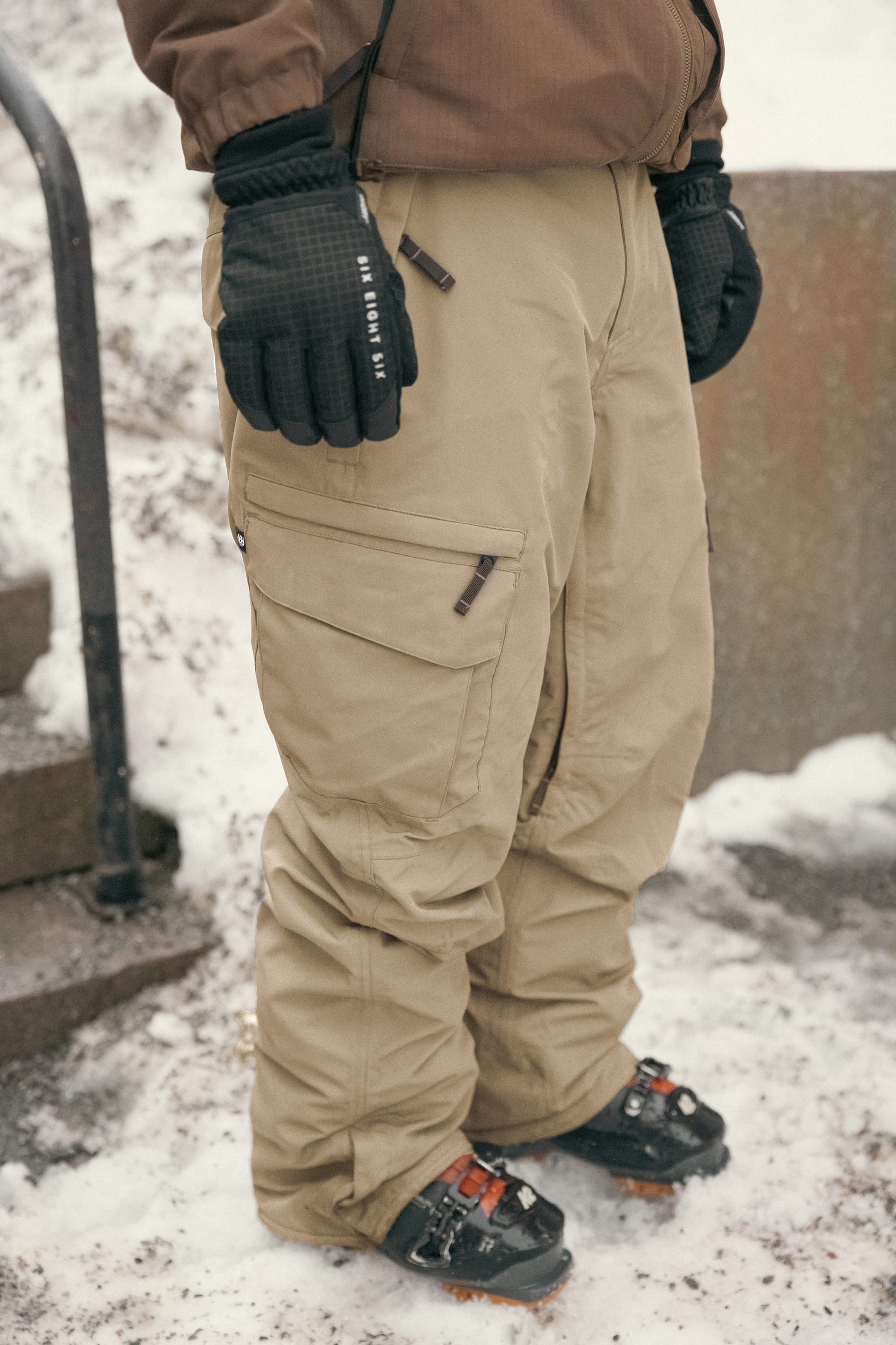 A person wearing 686 Men’s SMARTY® 3-in-1 Cargo Pant snow pants, black gloves, and crampon-fitted boots is standing on snowy ground near stairs and a metal object.