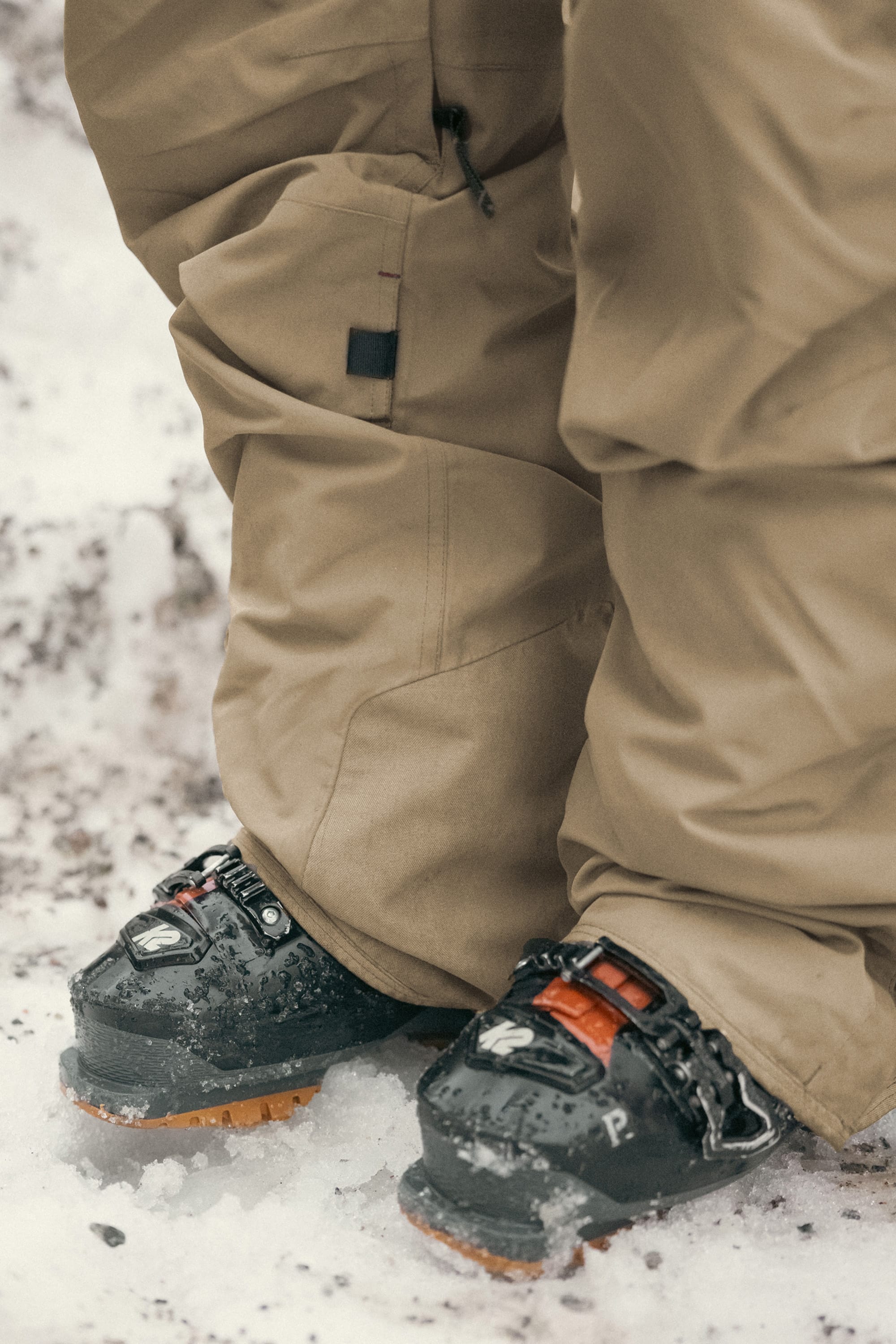 A person wearing 686 Men’s SMARTY® 3-in-1 Cargo Pant snow pants and black snowboard boots stands on snow-covered ground.