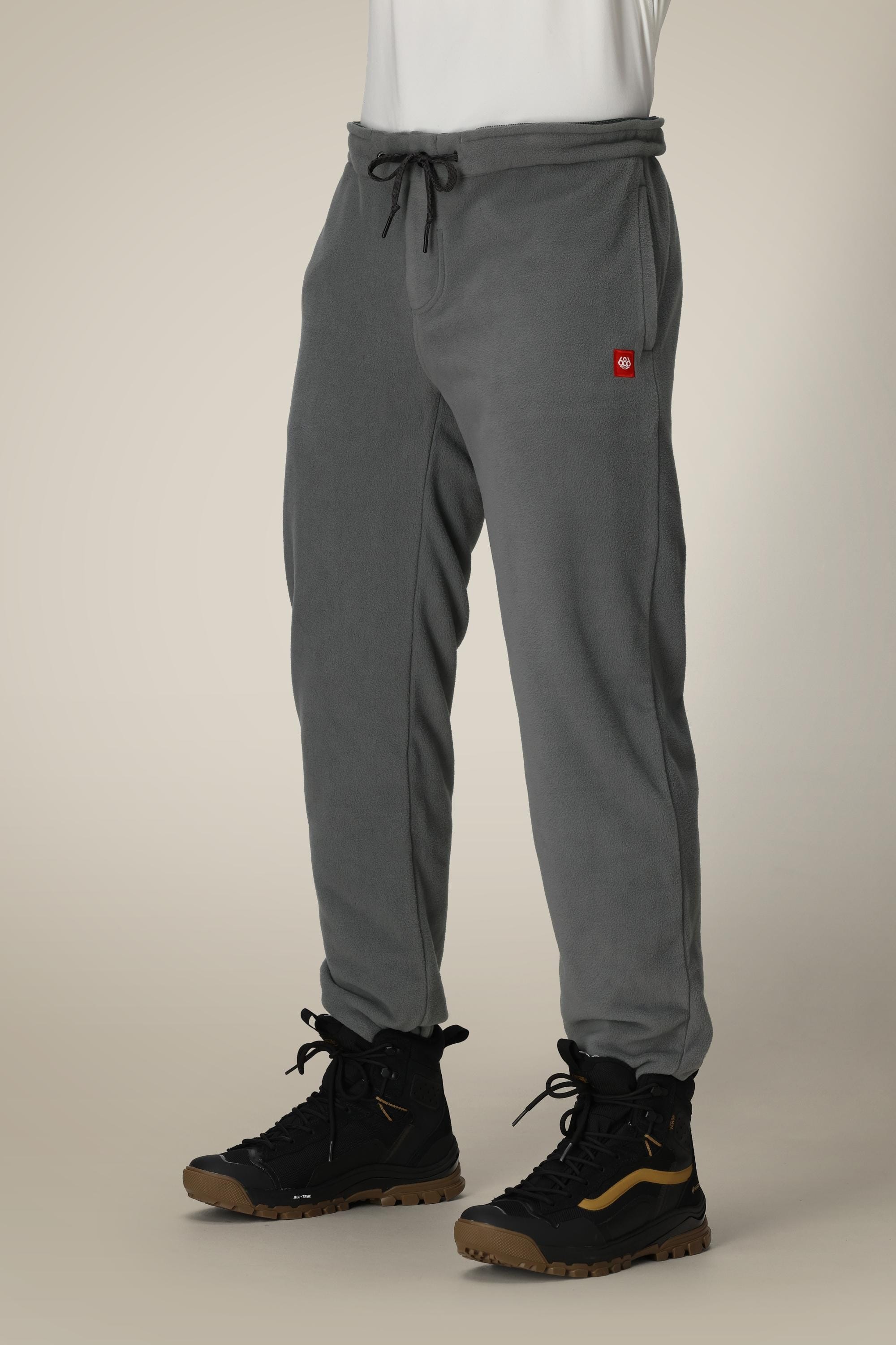 A person is wearing the 686 Men’s SMARTY® 3-in-1 Cargo Pant, featuring a gray fleece fabric with a drawstring waist and a small red square logo on the upper left leg, paired with black and brown hiking boots. The background is neutral beige.