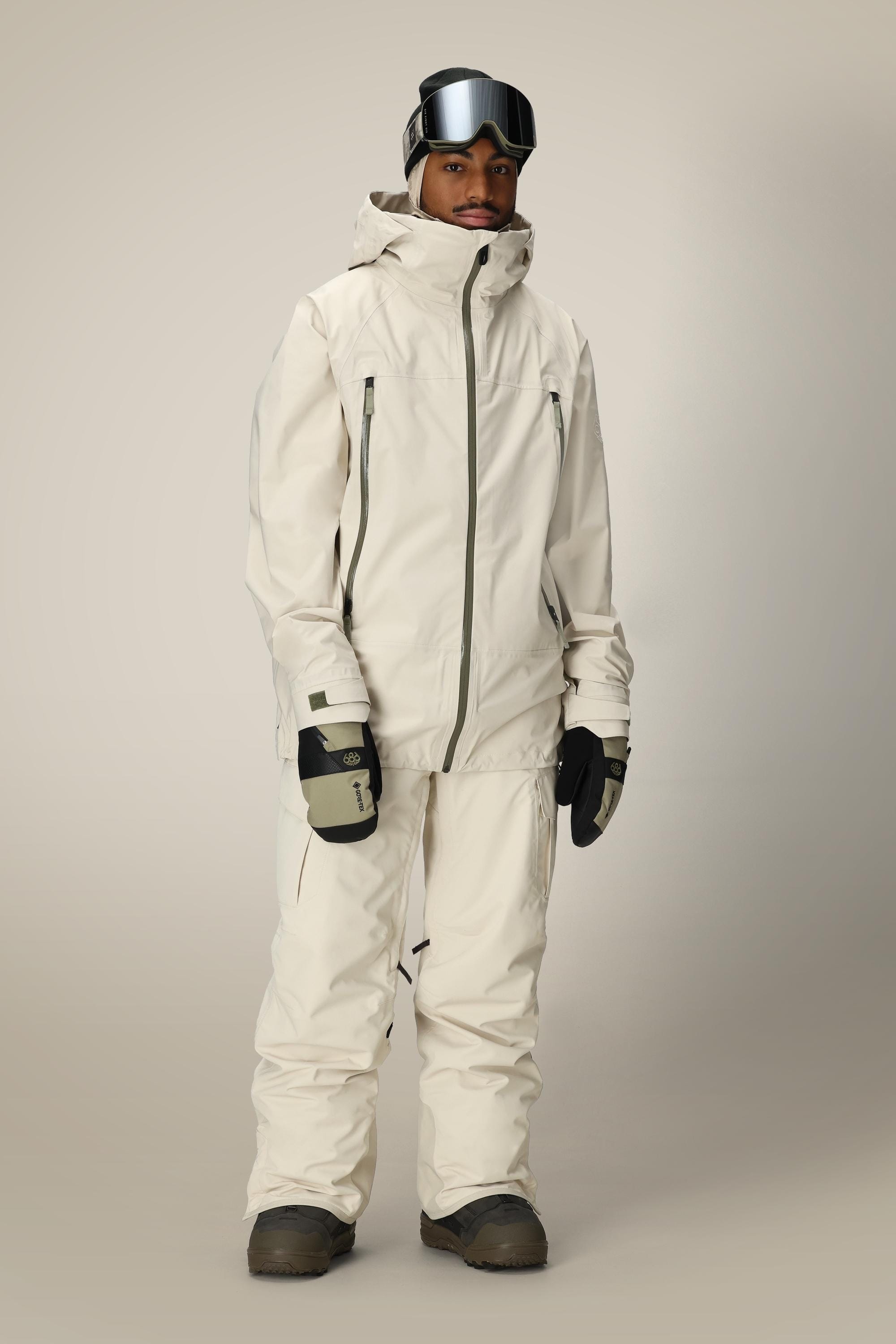 A person wears a cream ski jacket and 686 Men's SMARTY® 3-in-1 Cargo Pant by 686, featuring a removable sweatpant liner, plus black gloves, brown boots, and ski goggles, standing against a plain light background.