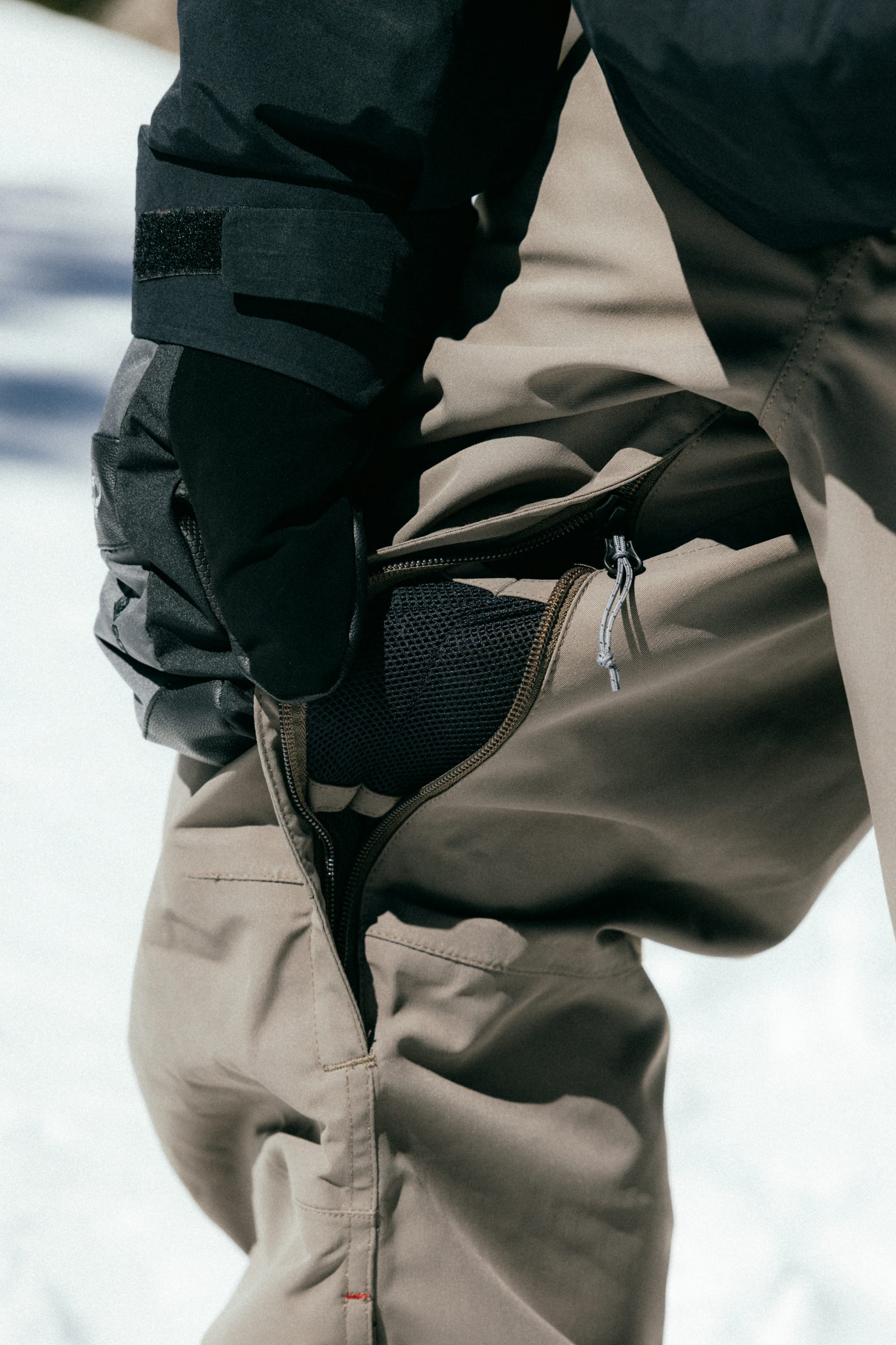 A person in 686 Men's SMARTY® 3-in-1 Cargo Pant stands on snow, unzipping a vent to reveal mesh, highlighting its versatile wear—thanks to the removable sweatpant liner from 686.