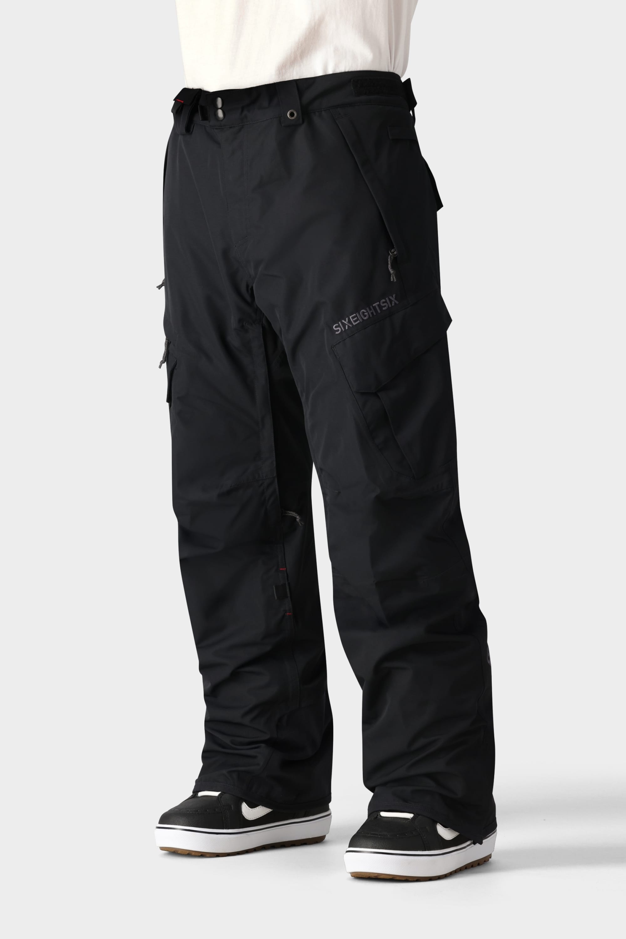 Only the lower half is visible: a person wearing 686 Men's SMARTY® 3-in-1 Cargo Pant—loose-fitting black cargos with multiple pockets and a removable sweatpant liner—paired with black-and-white sneakers against a light gray background.