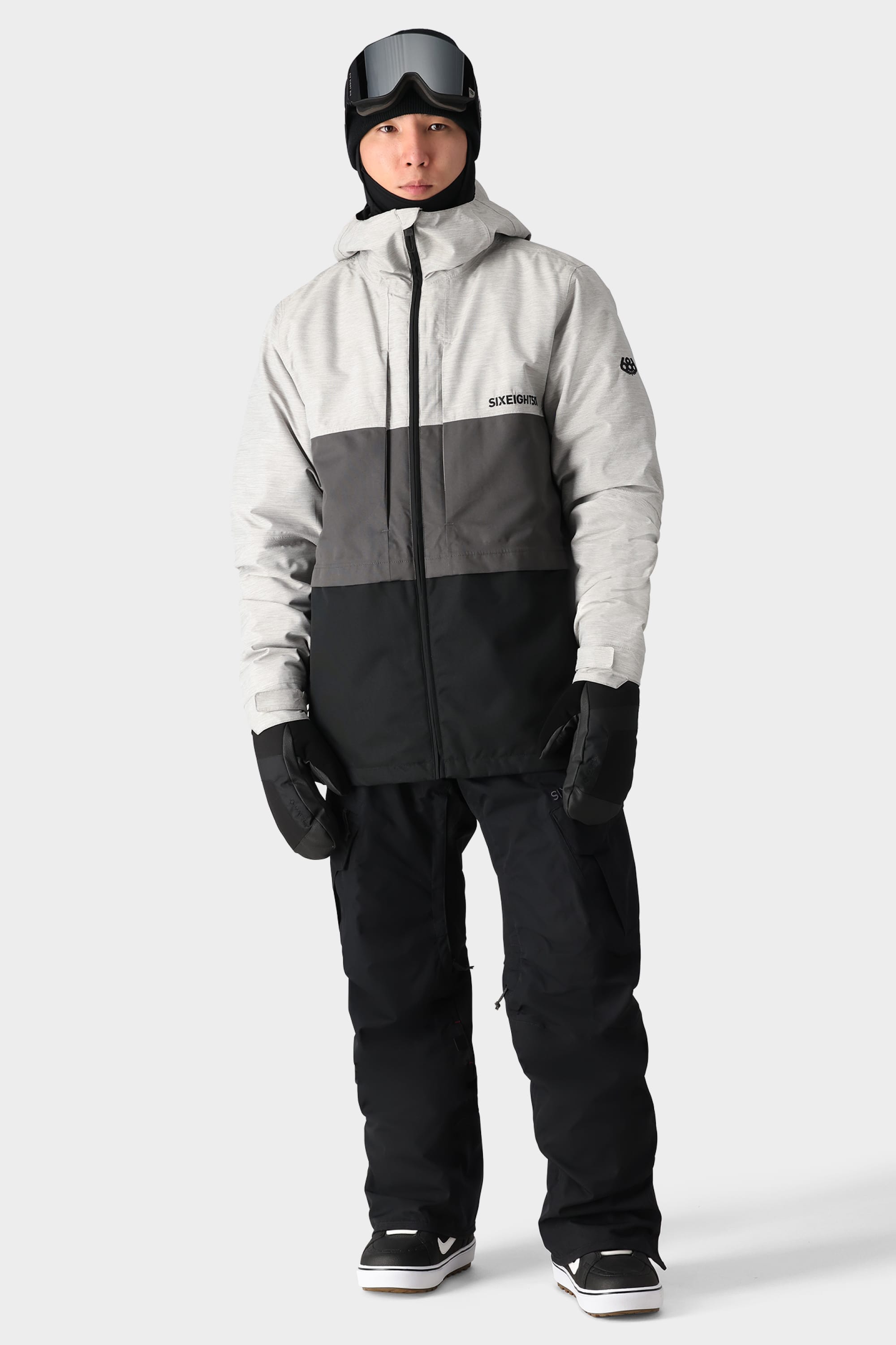 A person wearing 686 Mens SMARTY 3-in-1 Cargo Pant by 686, a gray and black winter jacket, black gloves, cargo-style boots, a black helmet, and ski goggles stands against a plain light gray background.