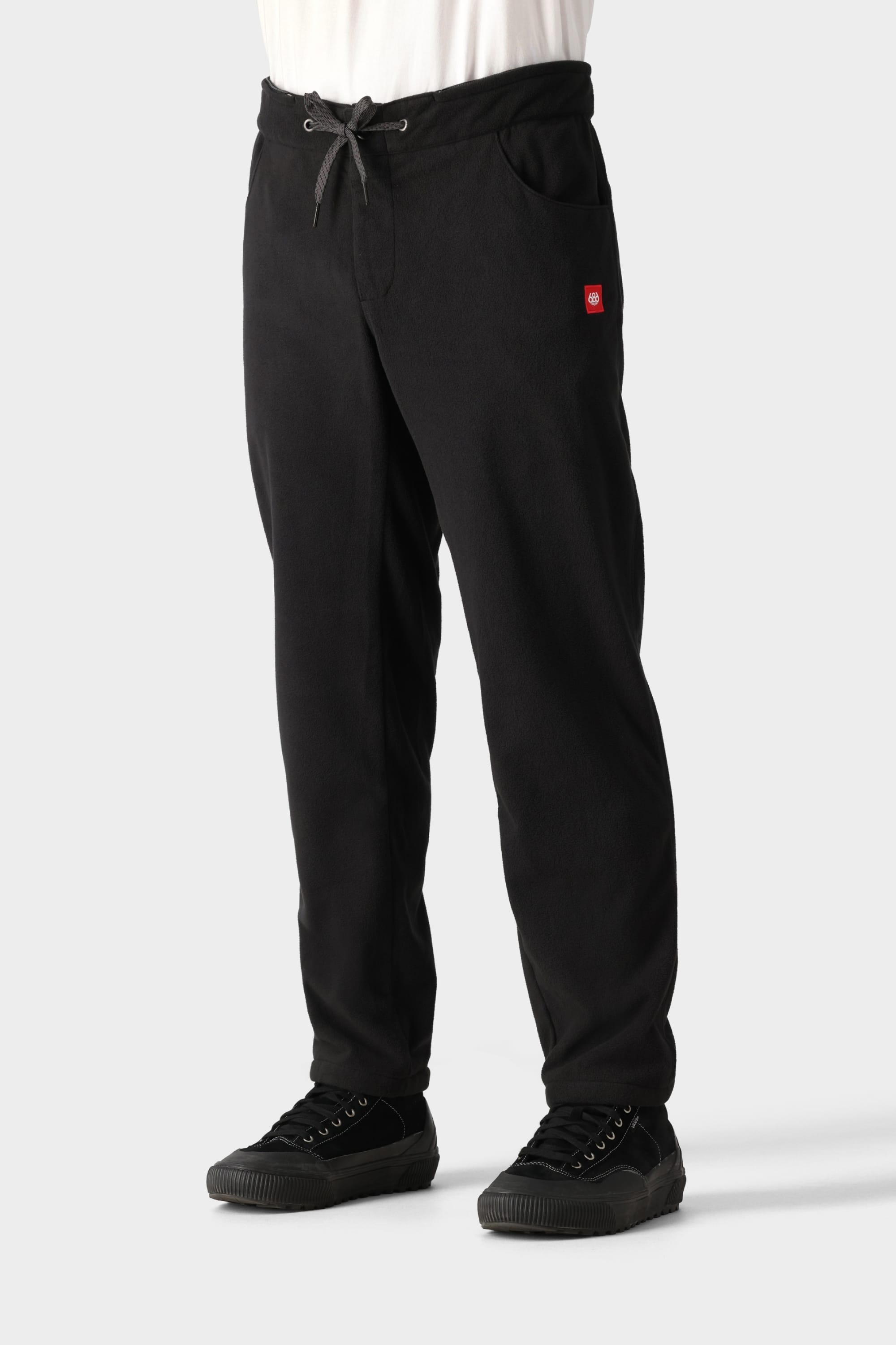 A person is wearing the 686 Men’s SMARTY® 3-in-1 Cargo Pant by 686, featuring a small red rectangular logo on the right side near the pocket, paired with black sneakers and a white shirt. The pants have drawstrings and pockets. The background is plain and light gray.