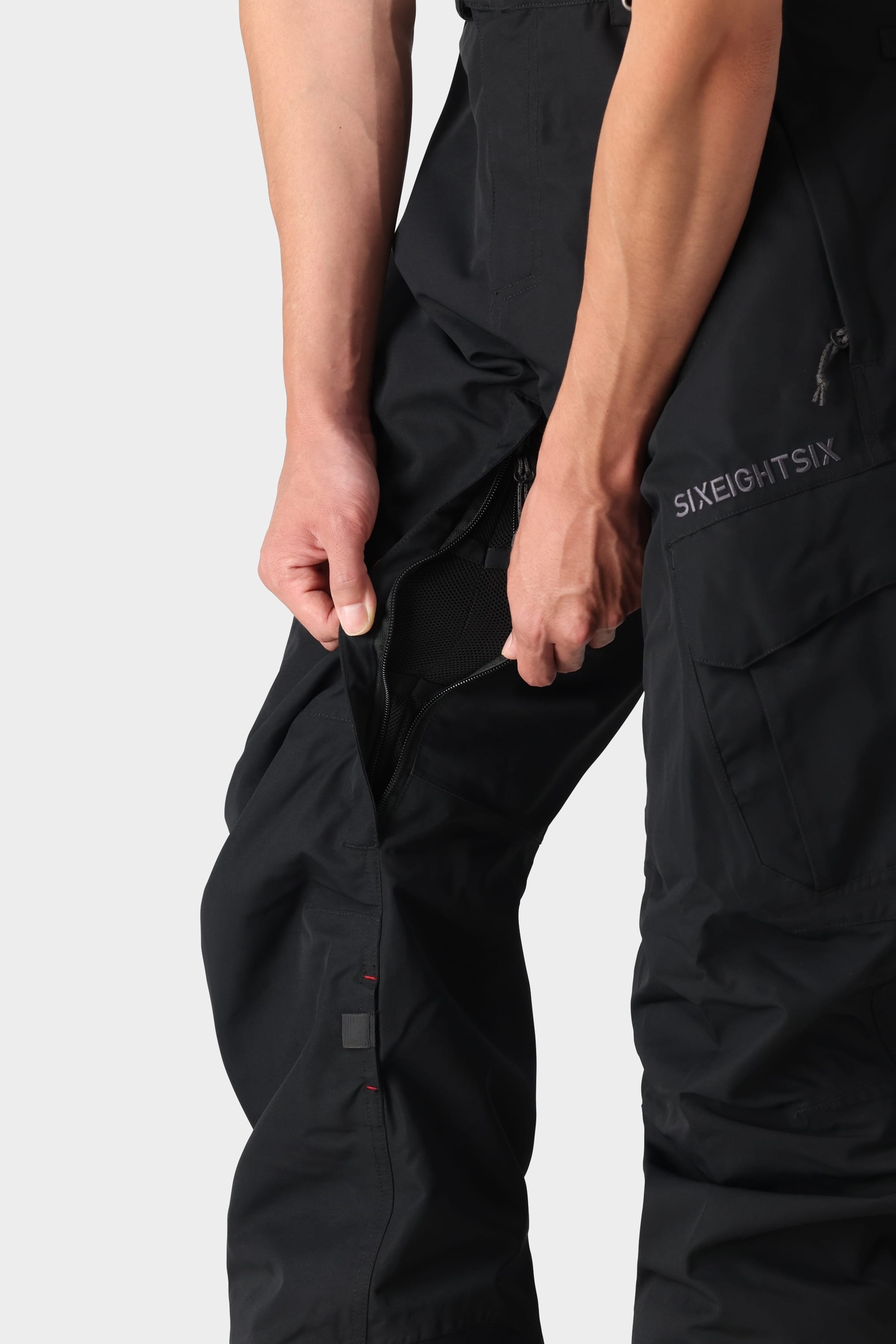 A person unzips the side vent of 686 Men’s SMARTY® 3-in-1 Cargo Pants, revealing mesh fabric underneath. Only the lower torso and hands are visible against a plain background.