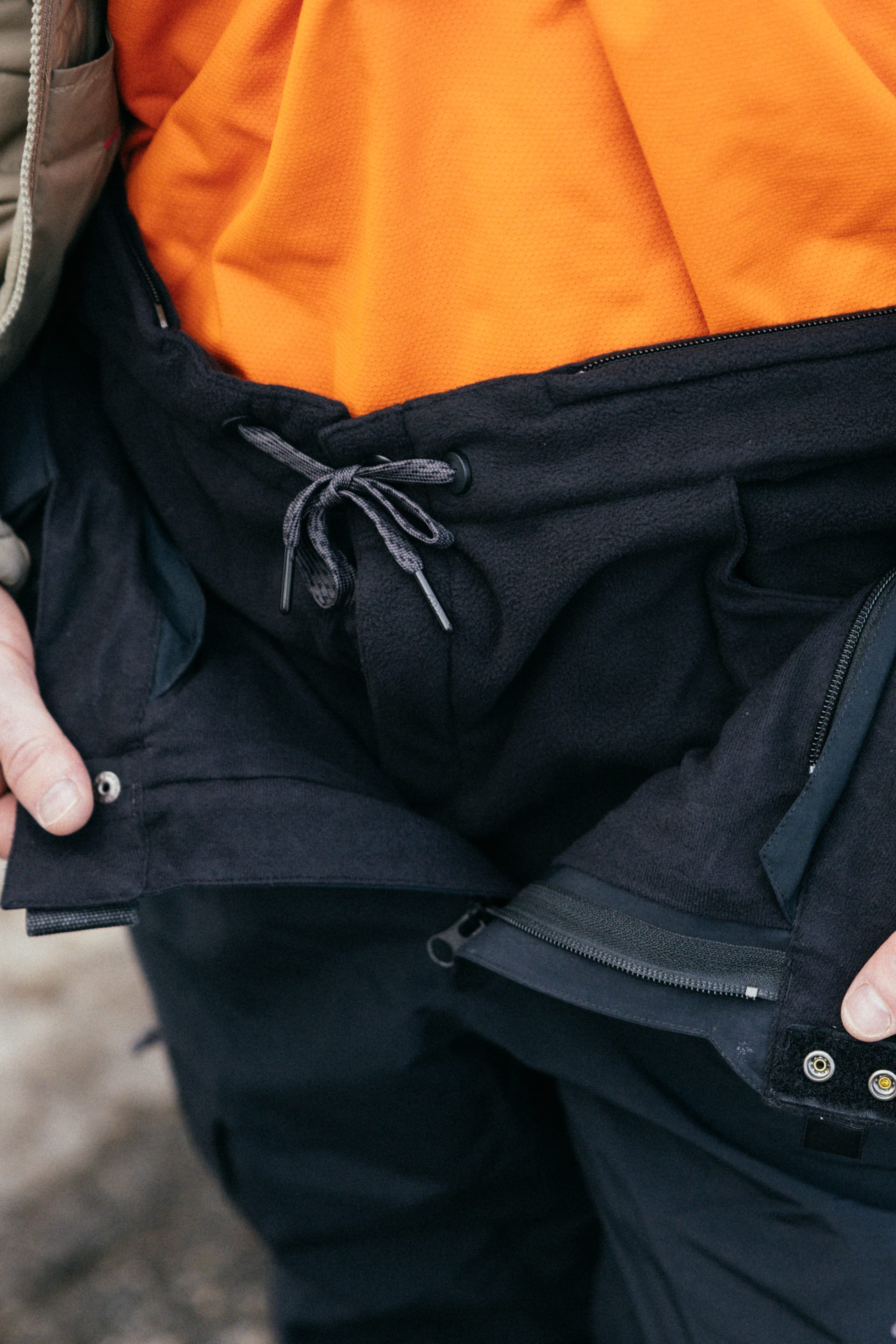 A close-up of a person wearing an orange shirt and the 686 Men’s SMARTY® 3-in-1 Cargo Pant in black, featuring a drawstring waist and unzipped to reveal layers of outdoor clothing.