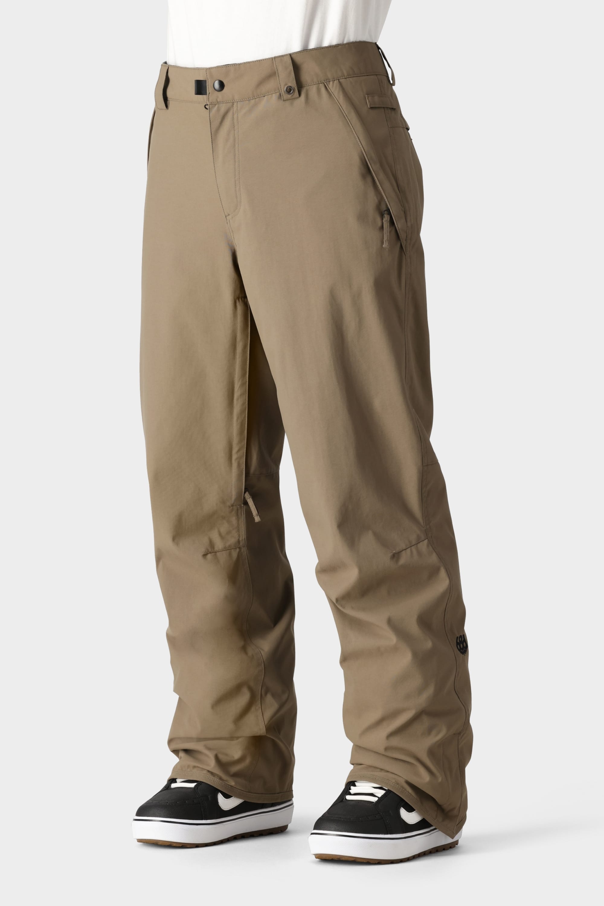 A person wears 686 Mens Standard Shell Pants in light brown, featuring 10k waterproofing, zippered pockets, and a relaxed fit, paired with black sneakers with white soles against a plain light gray background.