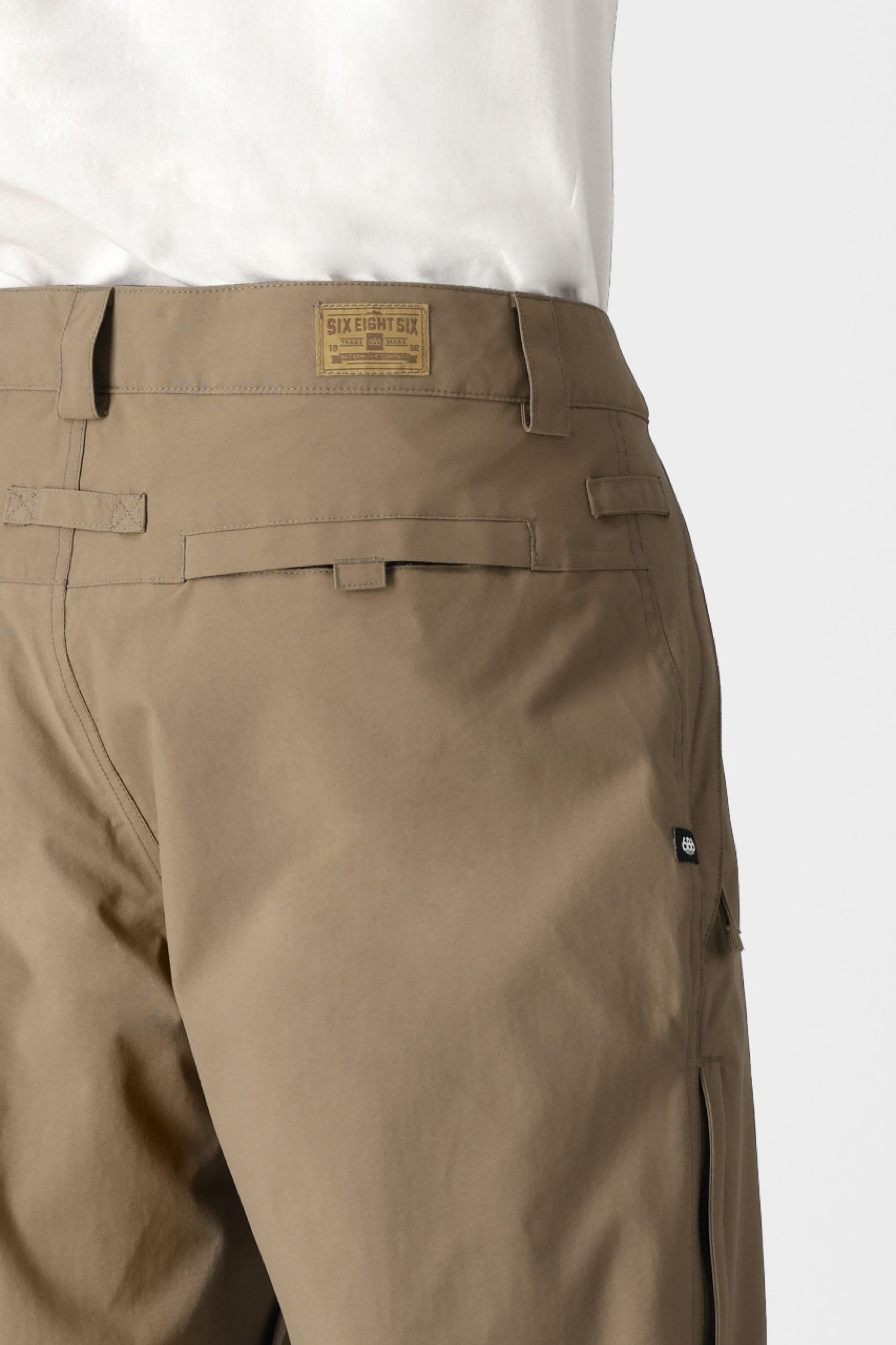 A close-up shows the back and side of a person wearing 686 Mens Standard Shell Pants with secure pockets and a visible 686 label on the waistband, paired with a white top.