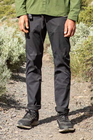 A person stands on a rocky trail wearing 686 Men's Everywhere® Pant - Slim Fit, black sneakers, and a green jacket. Their hands are relaxed by their sides, with green shrubs visible in the background.