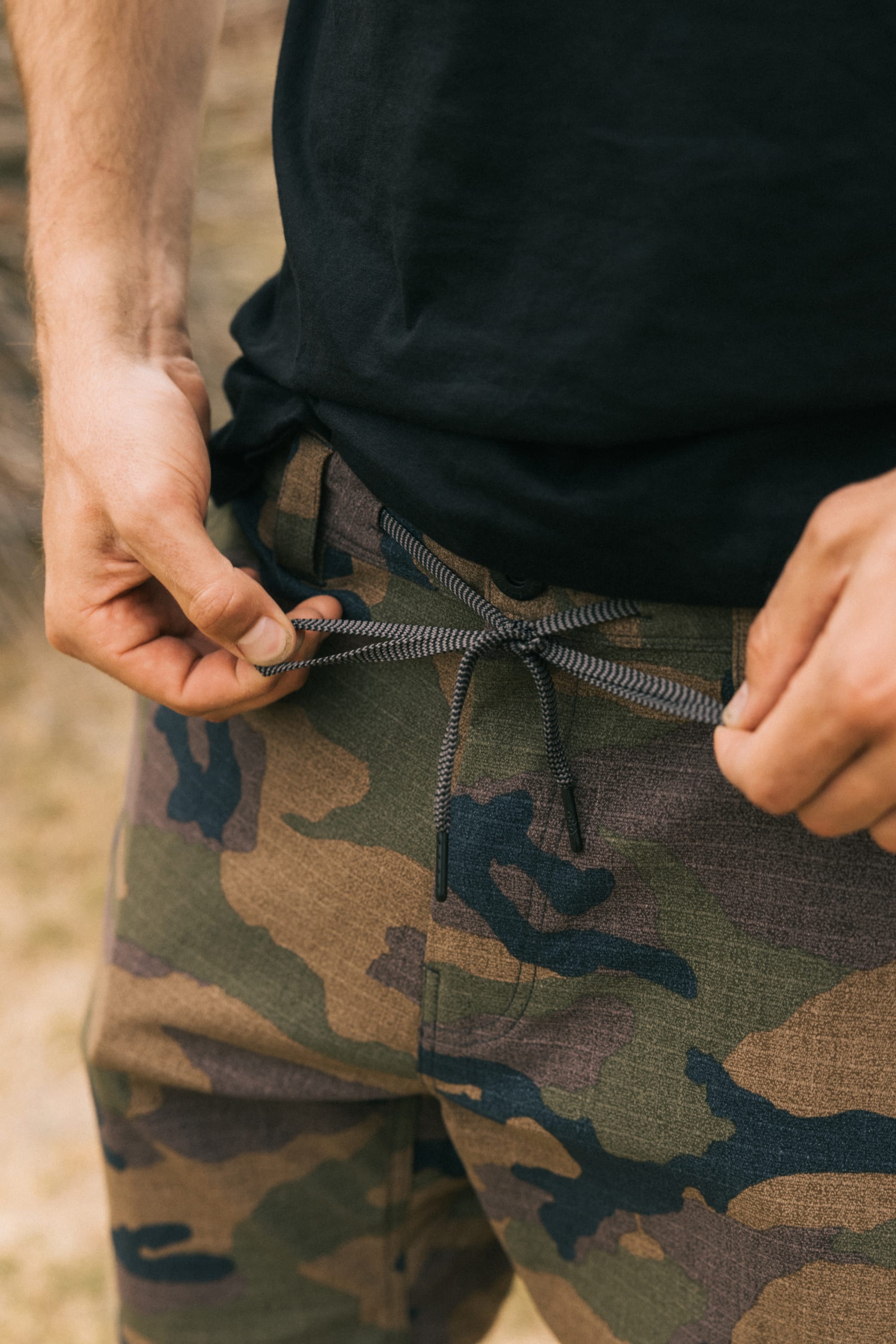 A person, visible from the waist down, ties the drawstring on breathable, stain-resistant 686 Men's Everywhere® Pant - Slim Fit camouflage pants while wearing a black shirt.