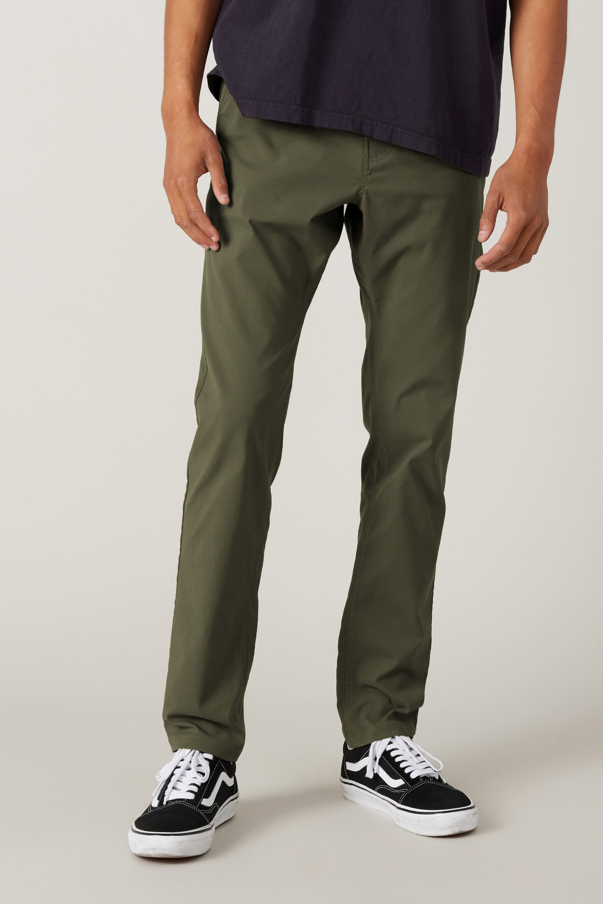 A person wearing 686 Men's Everywhere® Pant - Slim Fit in olive green with black and white sneakers and a dark t-shirt stands against a plain light background. The image shows the outfit cropped from the shoulders down.
