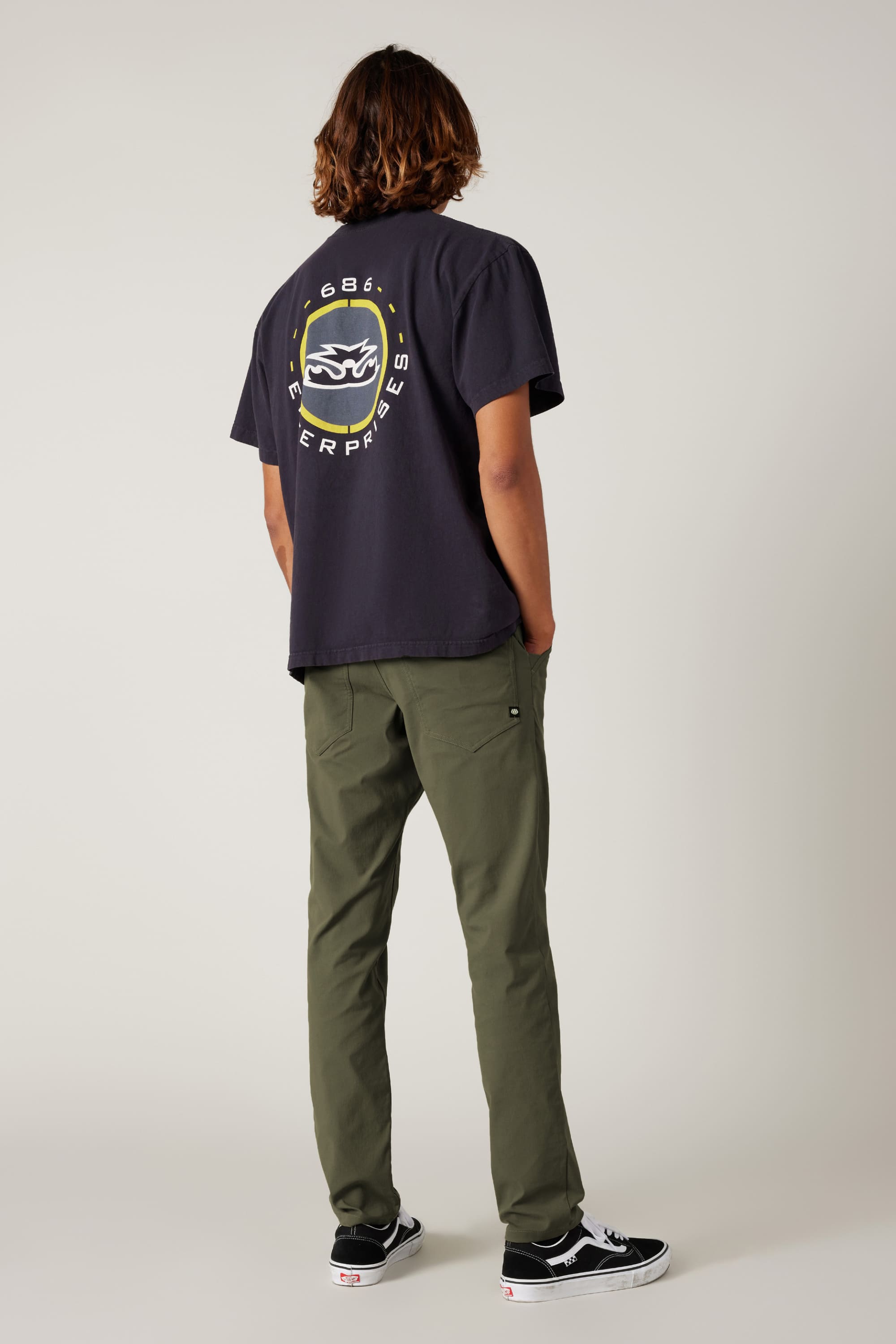 A person with shoulder-length hair faces away, wearing a black T-shirt, 686 Men's Everywhere® Pant - Slim Fit in olive green, and black Vans sneakers. The background is plain and light-colored.