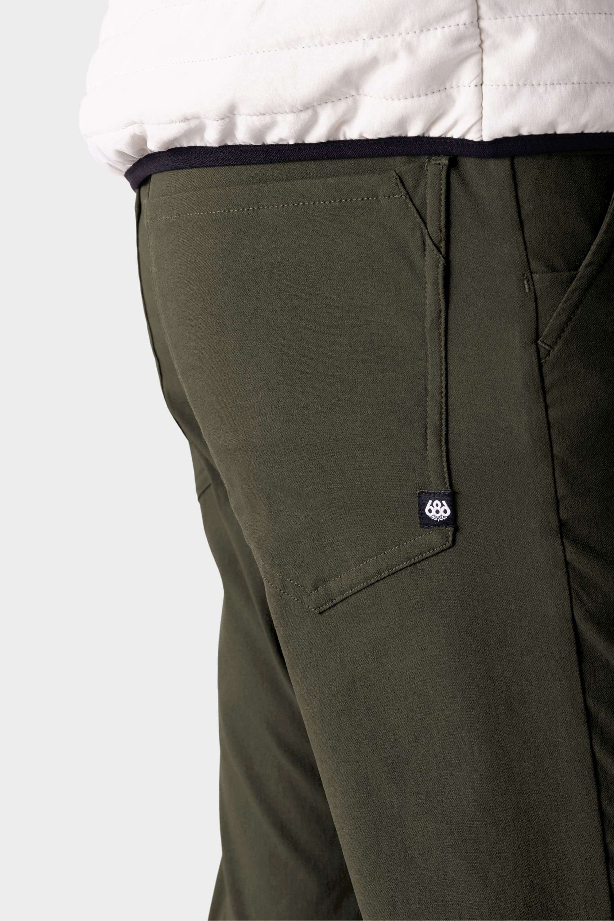 Close-up of a person wearing the 686 Men's Everywhere® Pant - Slim Fit in olive green, featuring a small pocket tag and detailed stitching. The image highlights the stain-resistant, breathable design paired with a white jacket.