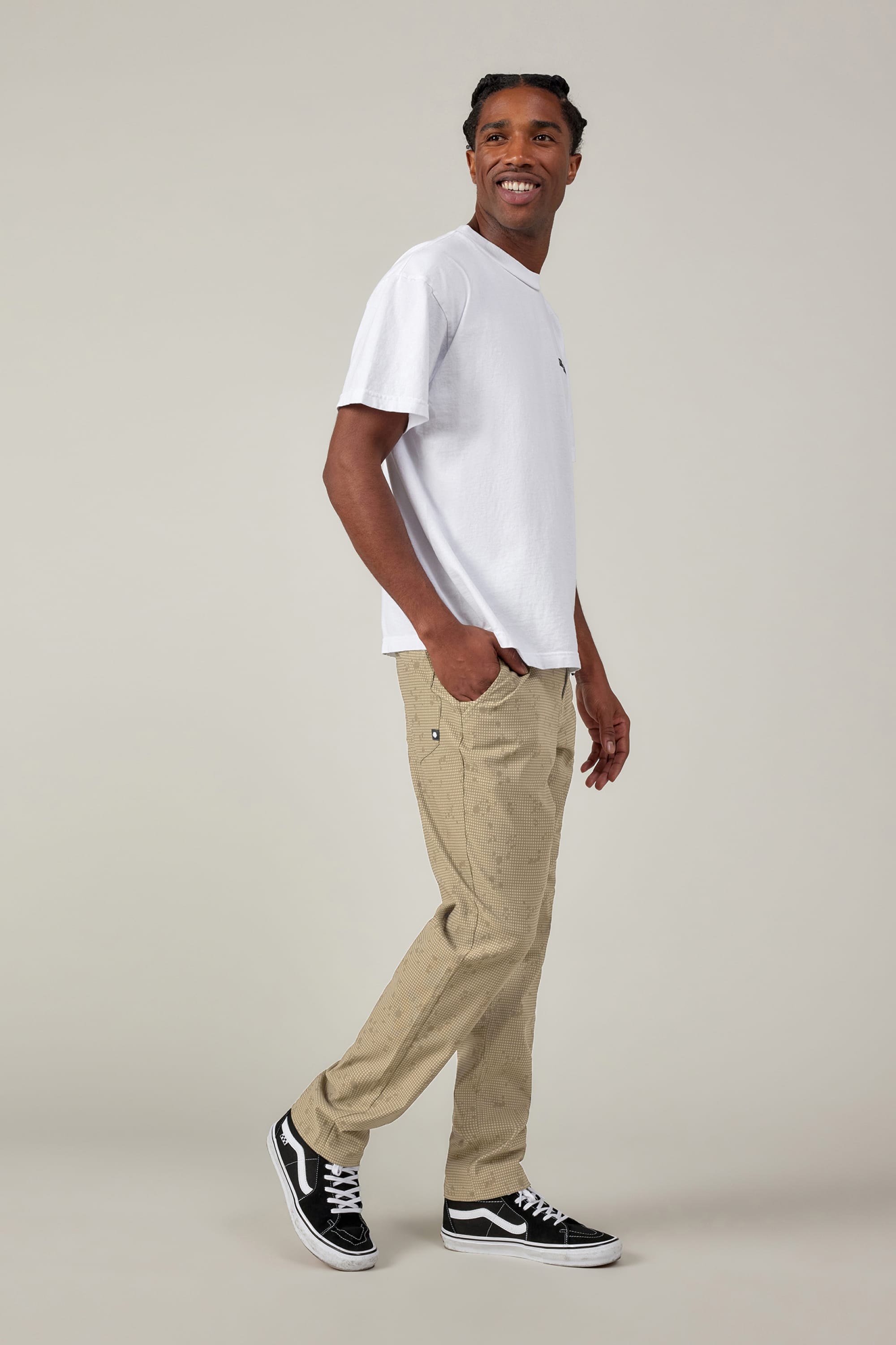 A man stands sideways, looking over his shoulder and smiling. He wears a white t-shirt, 686 Men's Everywhere® Pant - Slim Fit from 686 with subtle patterns, and black Vans sneakers against a plain light background.