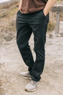 A person stands outdoors on a dirt path wearing 686 Men's Everywhere® Pant - Slim Fit by 686, paired with gray sneakers and a brown shirt. The background is blurred with brush and a trail sign visible. Hands are in pockets.