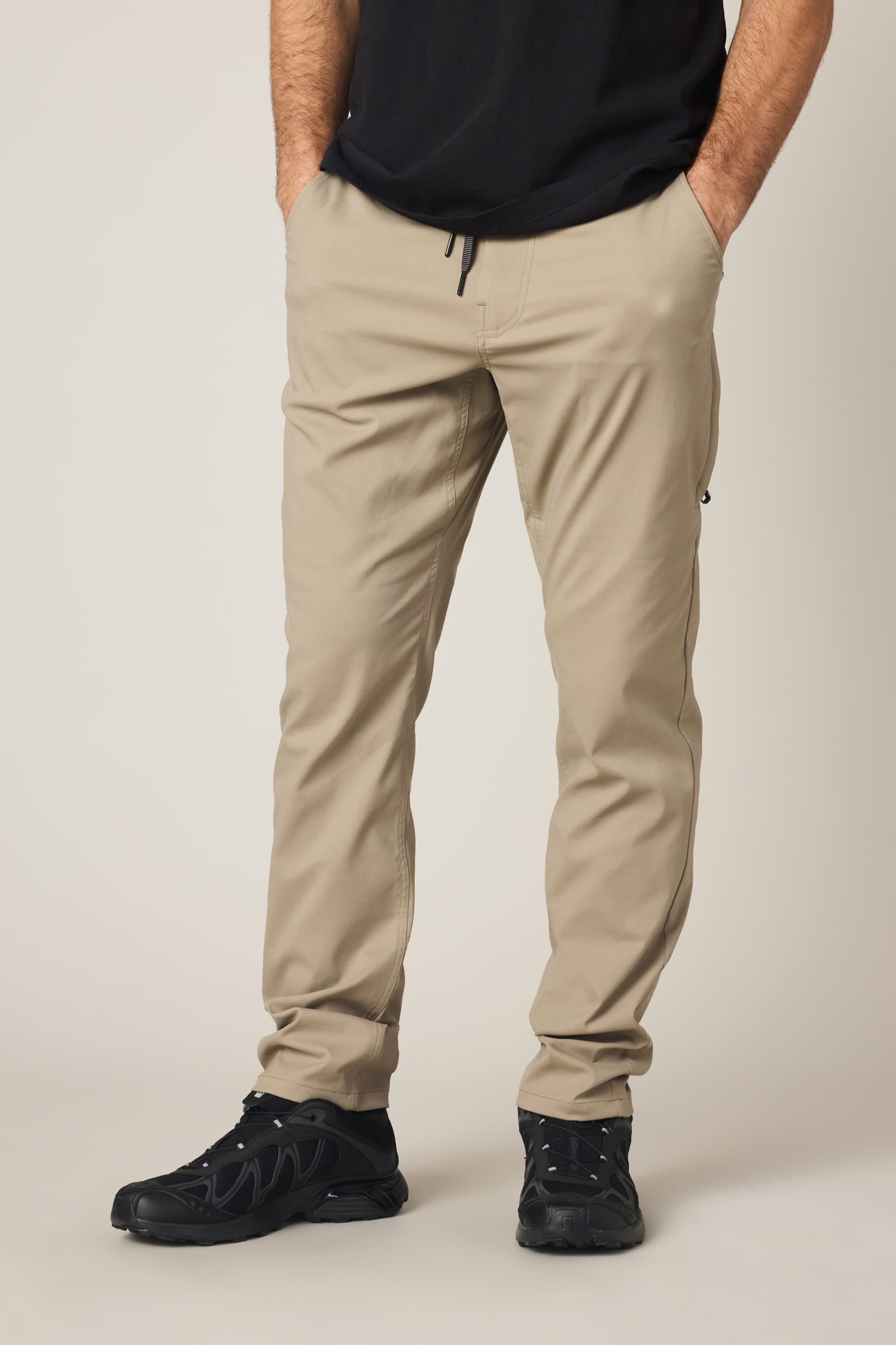 A person wearing 686 Men's Everywhere® Pant - Slim Fit in beige, paired with a black shirt and black sneakers, stands against a plain light background with hands in pockets.