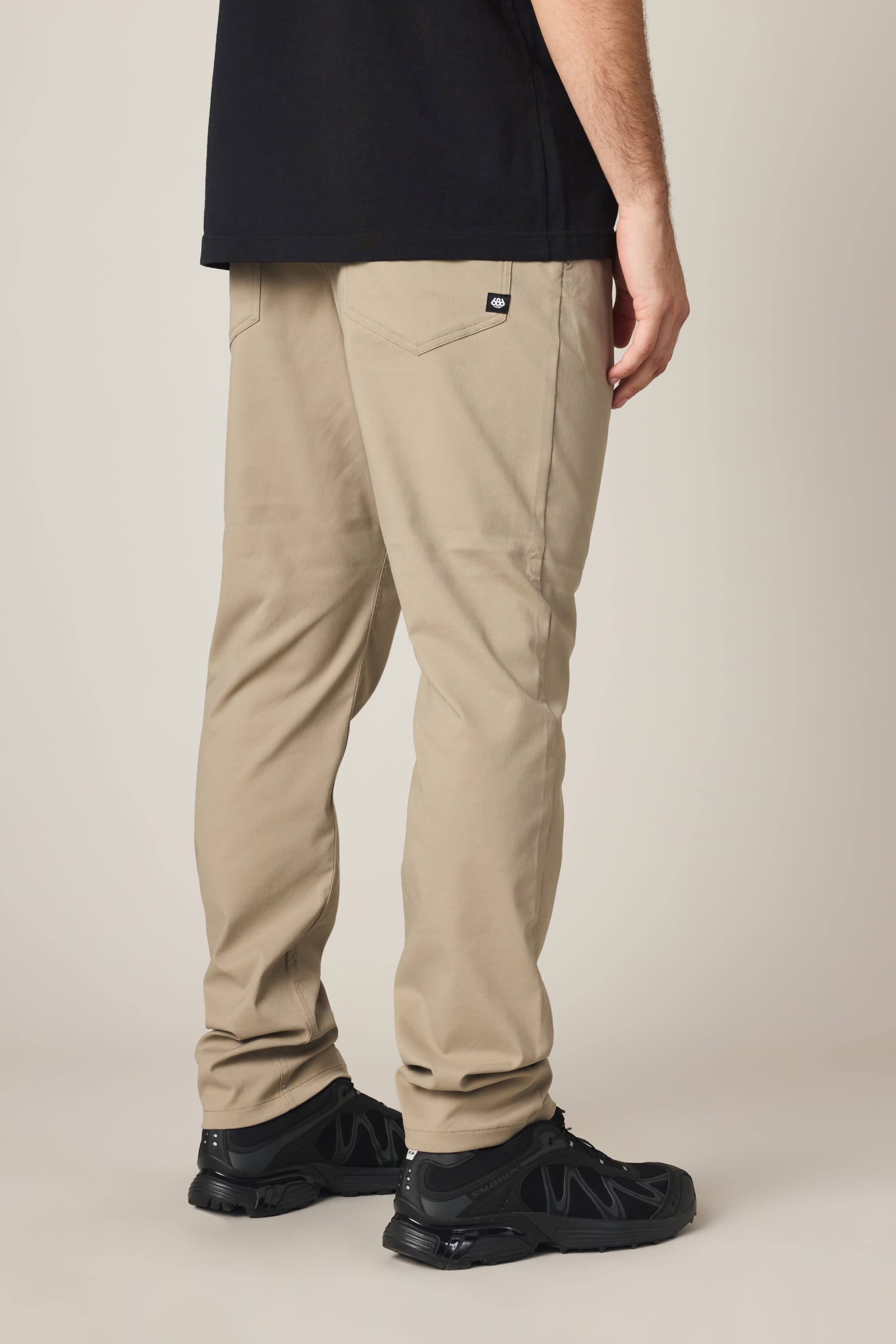 A person wearing 686 Men's Everywhere® Pant - Slim Fit in beige, paired with a black t-shirt and black sneakers, is standing with their back to the camera against a neutral background.