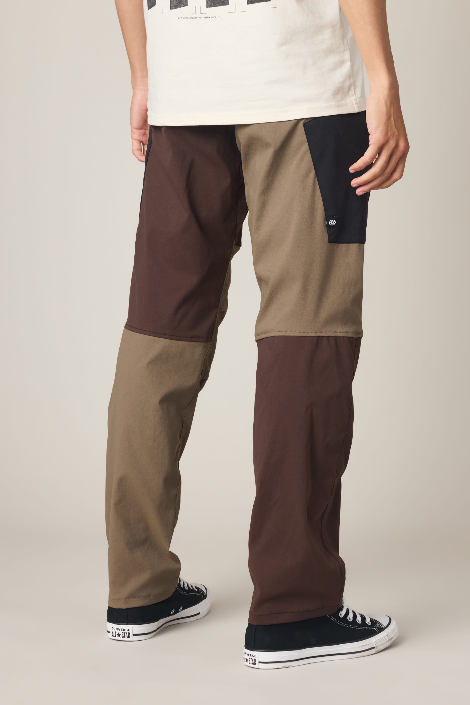 A person is shown from the waist down, facing away from the camera, wearing the 686 Men's Anything Cargo Pant - Relaxed Fit by 686. The technical cargo pants feature a color-blocked design in brown, olive, and black with 4-way stretch fabric and multiple 13-pocket detailing. Styled with black Converse sneakers, they stand on a neutral background.
