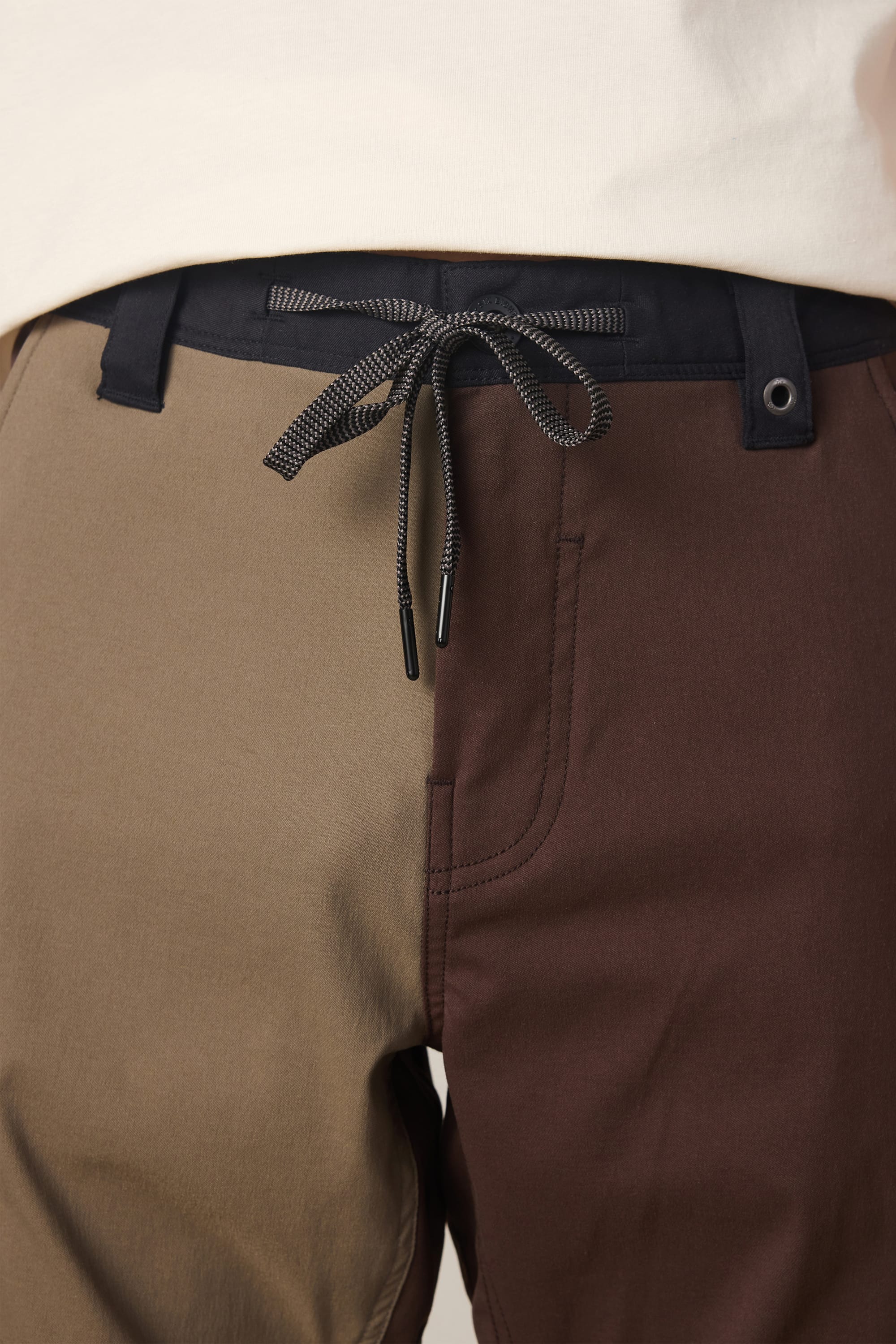 Close-up of a person wearing the 686 Men's Anything Cargo Pant - Relaxed Fit by 686, featuring a color-blocked design with one tan leg and one dark brown leg, a black drawstring tie at the waist, a visible front pocket, and made from 4-way stretch fabric for enhanced comfort.