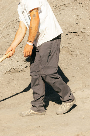 A person in a white shirt, 686 Men's Anything Cargo Pant - Relaxed Fit by 686, and checkered slip-on shoes stands on dusty ground holding a wooden stick. The person's face is not visible.