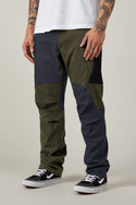 A person in 686 Men's Anything Cargo Pant - Relaxed Fit (olive green/dark gray, 13 pockets) with a white tee, black skate shoes, and tattooed arms stands against a plain background.