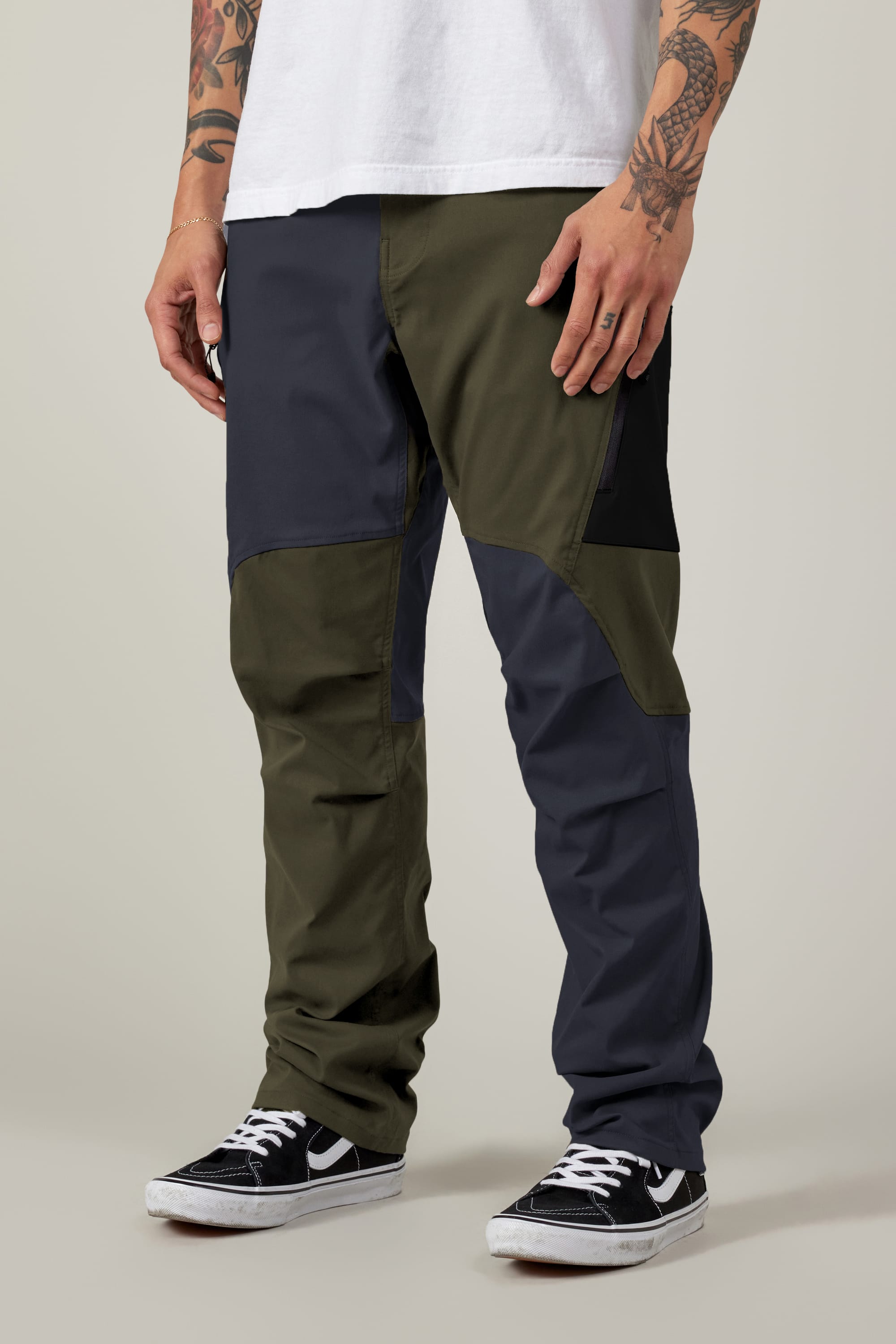 A person in 686 Men's Anything Cargo Pant - Relaxed Fit (olive green/dark gray, 13 pockets) with a white tee, black skate shoes, and tattooed arms stands against a plain background.