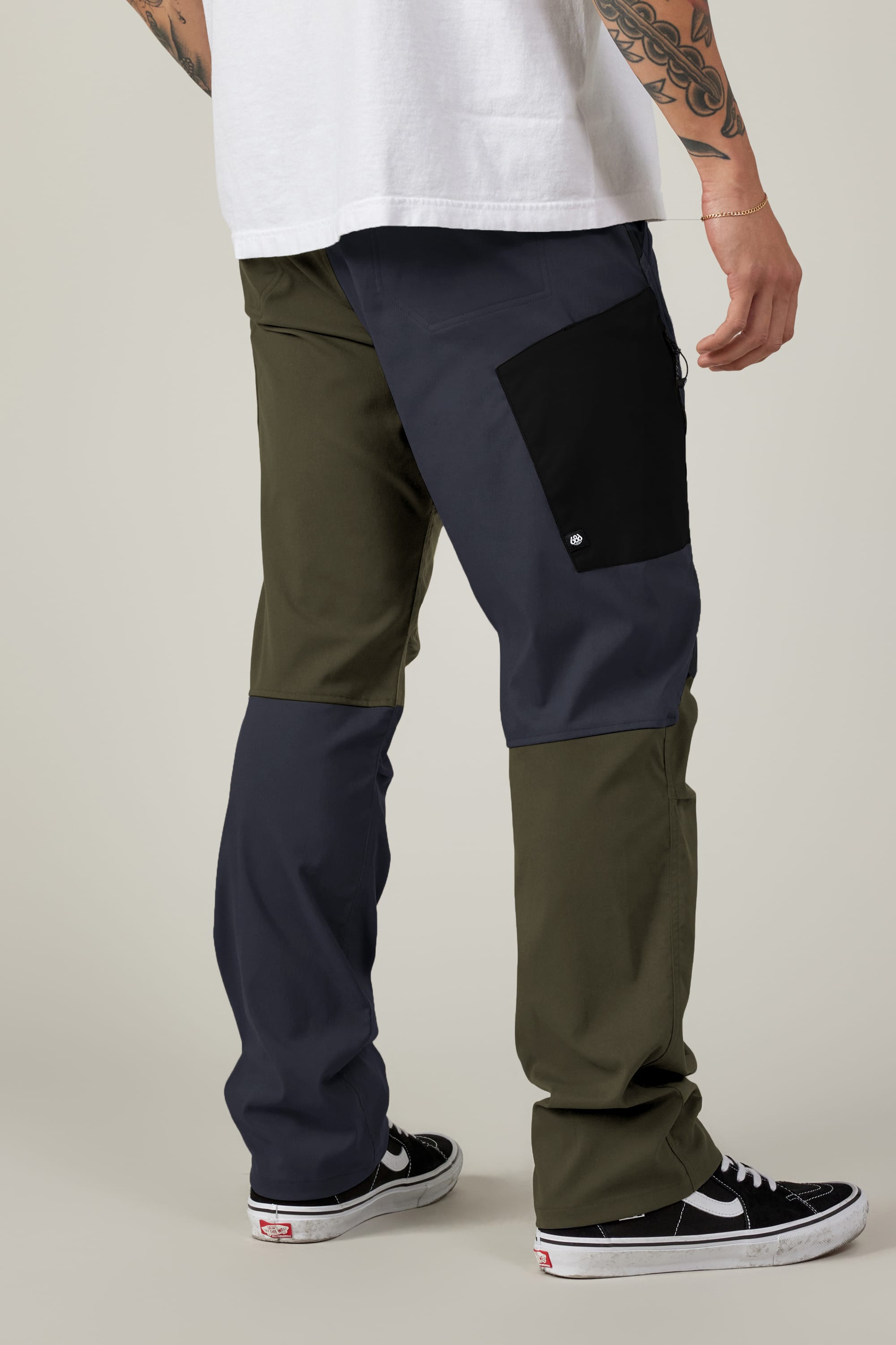 A person wears 686 Men's Anything Cargo Pant - Relaxed Fit by 686 in olive green, navy, and black, paired with a white t-shirt and black Vans sneakers. Tattoos cover their right arm. The scene has a plain light-colored background.