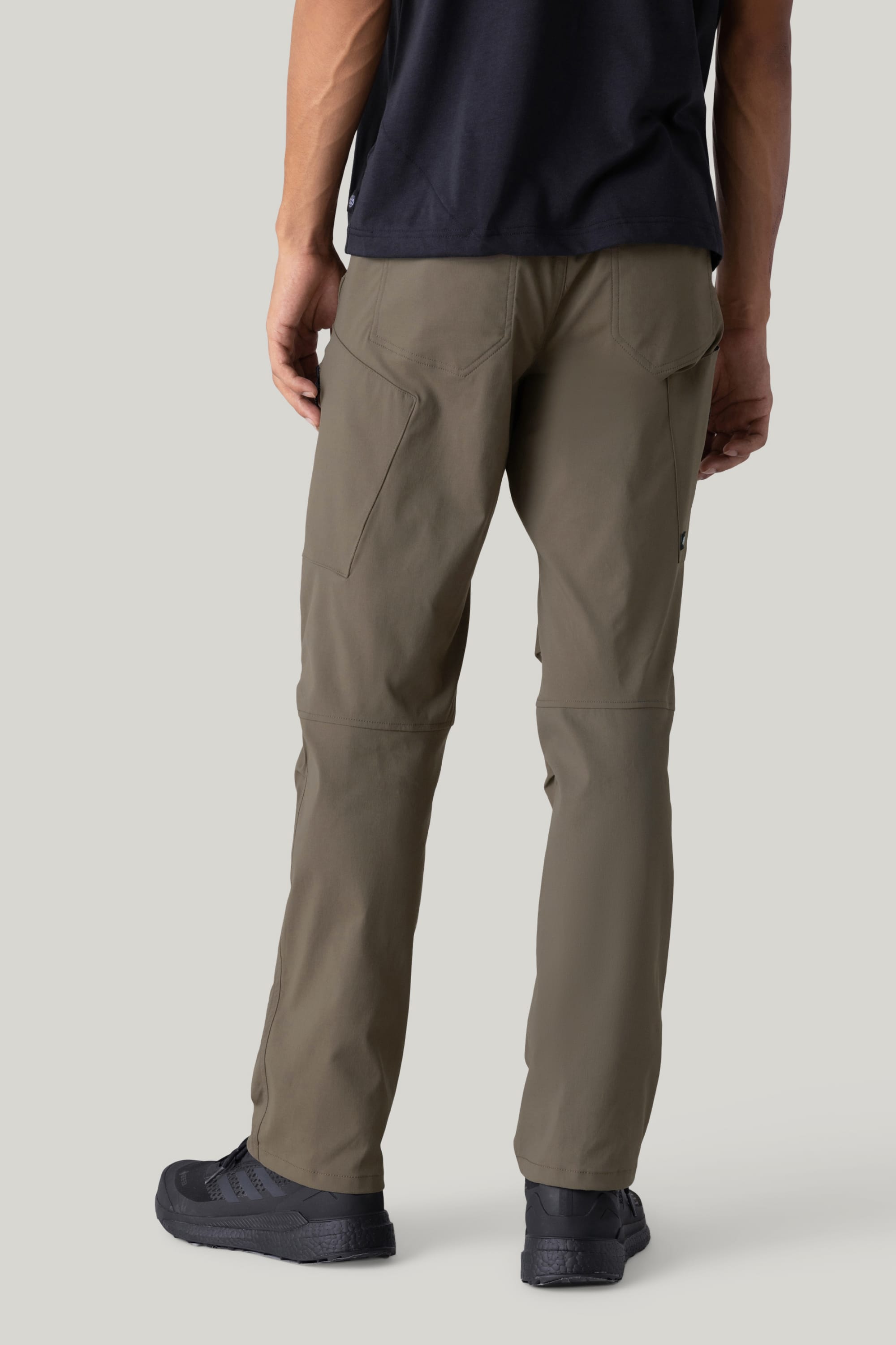 A person is shown from the back wearing 686 Men's Anything Cargo Pant - Relaxed Fit by 686 in olive green, paired with a black shirt and black athletic shoes, standing against a plain light gray background.