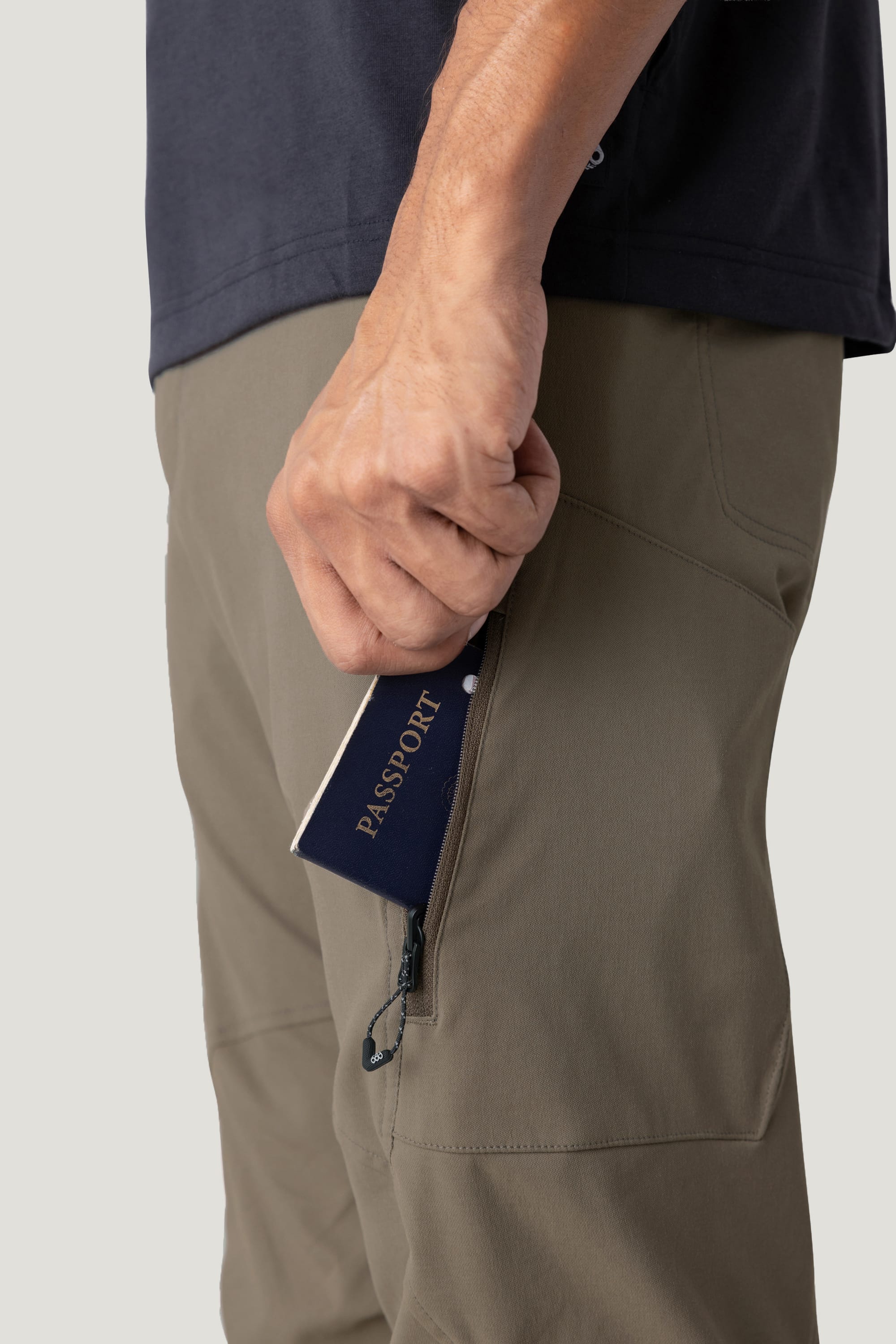 A hand places a navy blue passport into the zippered pocket of the 686 Men's Anything Cargo Pant - Relaxed Fit (olive green), with part of a dark shirt visible.