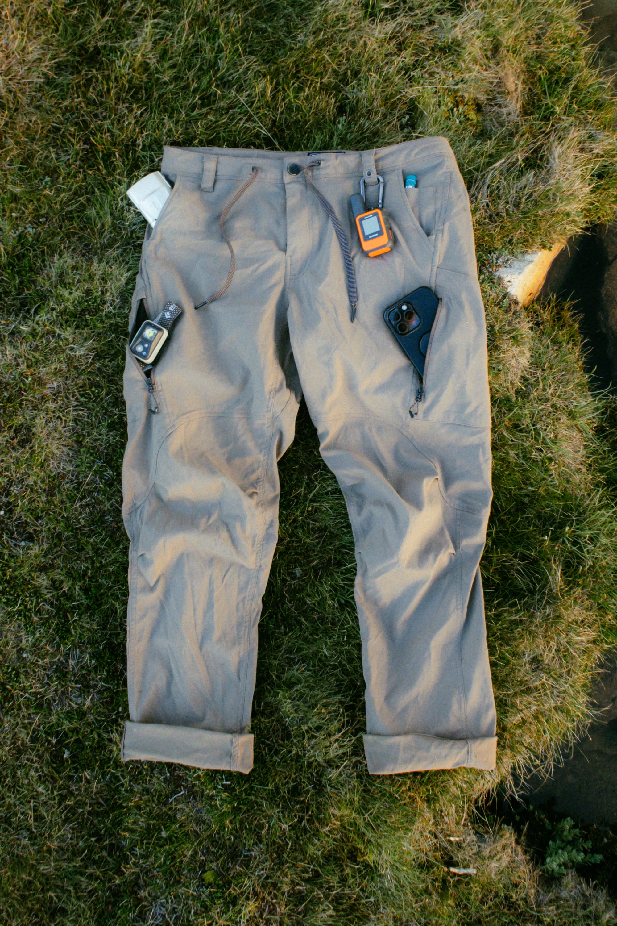 The 686 Men's Anything Cargo Pant - Relaxed Fit by 686 is shown on grass, its 13 pockets and waistband holding outdoor gear like phones, GPS devices, and a notepad.