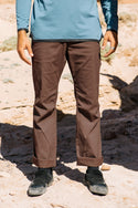 A person stands outdoors on rocky ground, wearing 686 Men's Everywhere® Pant - Relaxed Fit in brown, black shoes, and a long-sleeve blue shirt. The image is cropped at the torso to show only the lower half of the body.