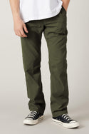 A person wearing 686 Men's Everywhere® Pant - Relaxed Fit in olive green stands against a plain light background. Only the lower half of their body, paired with a white T-shirt and black low-top sneakers with white soles, is visible.